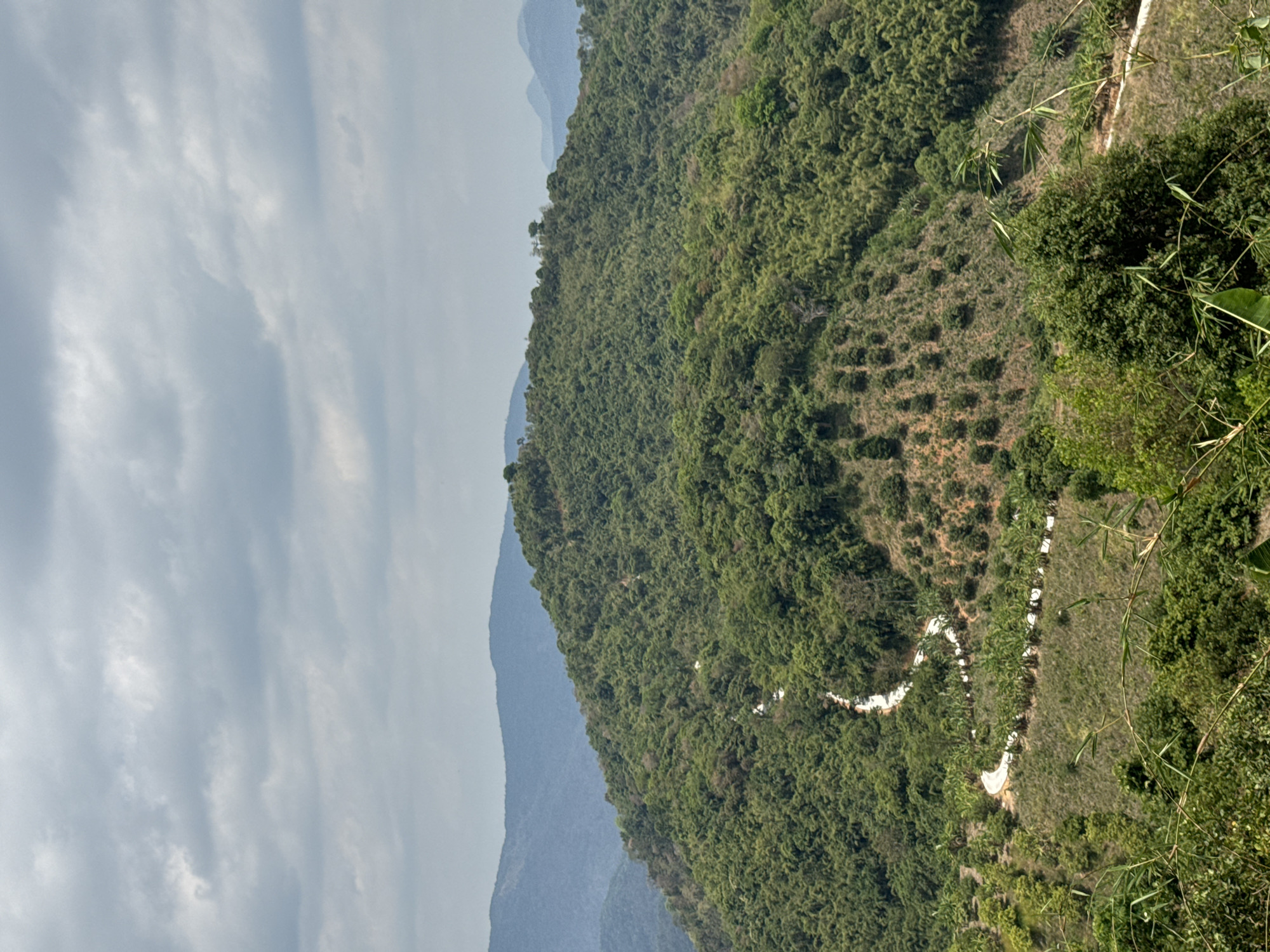 The image shows a landscape with green, tree-covered mountains under a cloudy sky, potentially taken in or near Son Dien, Vietnam. The mountain range extends from the foreground to the background. A winding, light-colored path or road is visible on the side of the mountain in the right side of the frame. The foreground contains some greenery and trees. In the background, there are distant, hazy mountain peaks.