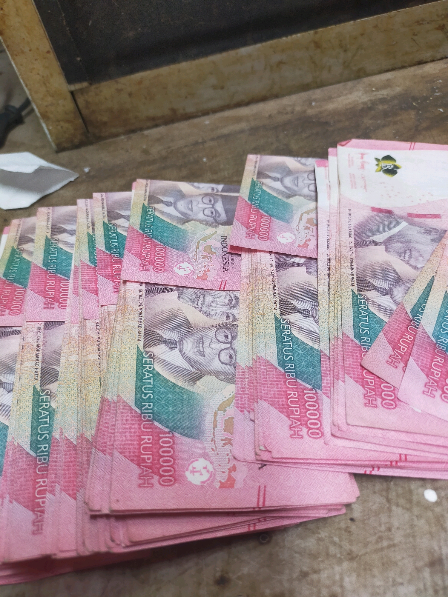 A pile of Indonesian Rupiah banknotes is spread out on a wooden surface. The banknotes are pink and feature portraits of prominent Indonesian figures. The denomination "100000" is visible on several notes, indicating they are 100,000 Rupiah bills. The bills are arranged in a fanned-out manner, with some overlapping others. The background is a rough, textured wooden surface, and a hint of a metal edge is visible at the top of the frame, suggesting the banknotes might be on a table or counter. The lighting is somewhat dim, casting soft shadows. The scene is devoid of any human presence or activity, presenting a still life composition of currency. The location is identified as Mojodanu, Indonesia.