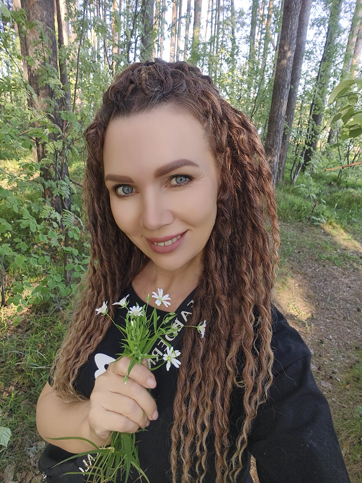 A woman is standing in a forest, holding a small bouquet of white flowers. She has blue eyes and long, curly brown hair. She is wearing a black shirt and appears to be looking directly at the camera. The background consists of trees and green foliage, suggesting a wooded area. The location of the scene is not specified.