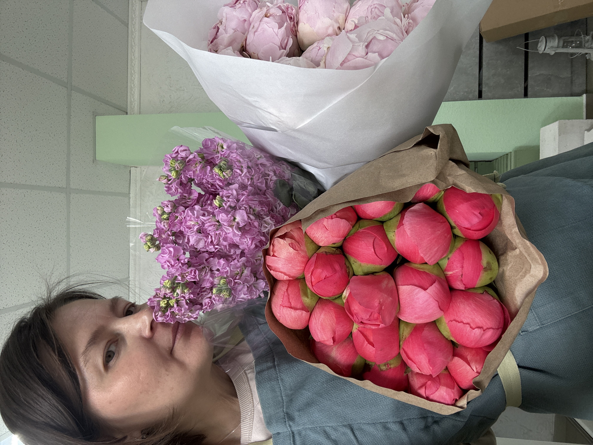The image shows a woman posing with three bouquets of flowers. One bouquet contains pink peonies wrapped in white paper. Another has purple stock flowers encased in plastic. The third bouquet features pink peonies in brown paper packaging. The woman has brown hair and is wearing a blue jacket. The location is indeterminate, and there is no evidence to suggest the image was taken in Izhevsk, Russia. The environment appears to be an indoor setting.