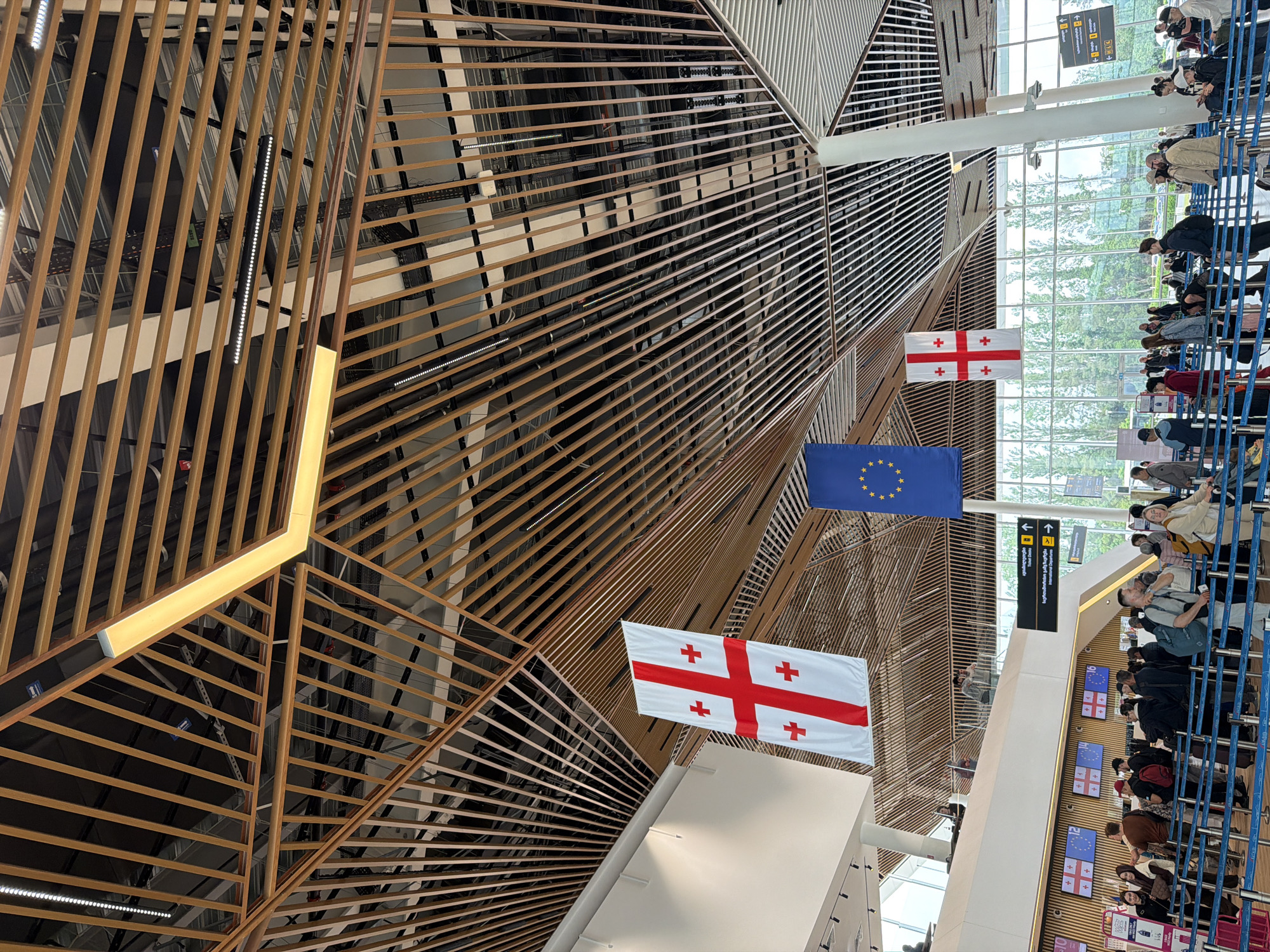 The interior of Kutaisi International Airport in Zeda Bashi, Georgia, displays the flags of Georgia and the European Union suspended from the ceiling. A waiting line of people, primarily seen from the side, is separated by blue metal barriers. Overhead signs direct passengers. The airport’s architectural ceiling features a pattern of wooden slats.