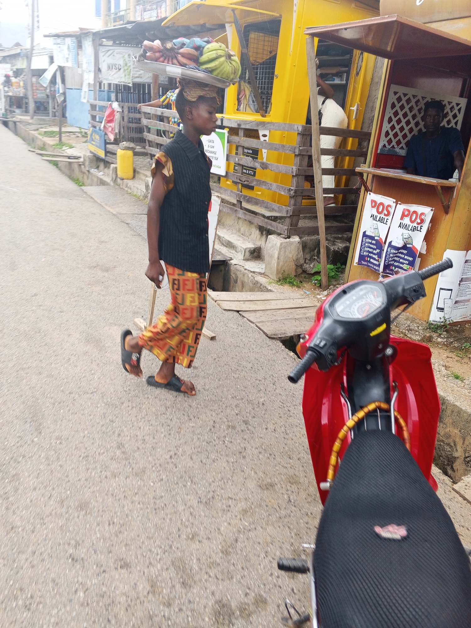 In Lokoja, Nigeria, a young person walks on a paved road, carrying a tray of bananas and other fruits on their head. The person wears a black short-sleeved shirt and brightly colored, patterned pants. They are walking past a yellow structure that appears to be a mobile shop or kiosk, from which a man in a blue shirt is visible inside. Two posters advertising "POS AVAILABLE" are affixed to the front of the kiosk. In the foreground, the seat and handlebars of a red motorcycle are visible. The environment suggests an outdoor marketplace or roadside setting, with other buildings and structures in the background.