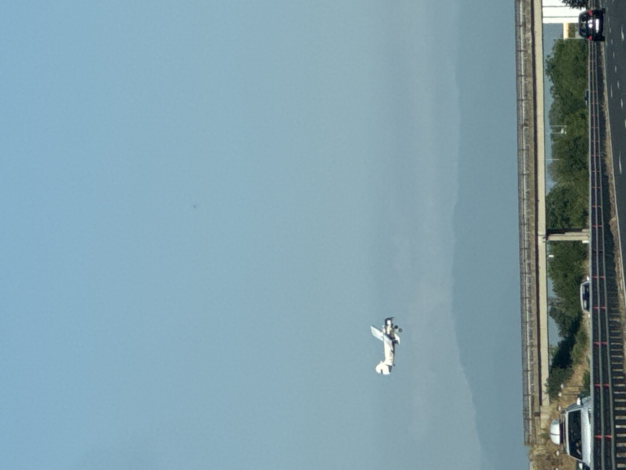 A biplane is in flight against a clear blue sky above a multi-lane highway. The airplane is white with dark accents and appears to be a propeller-driven aircraft. Below the plane, a highway overpass spans across the road. On the highway, several vehicles are visible, including a white SUV with a roof rack in the left lane and a dark-colored car in the right lane of the opposite direction. The environment suggests an outdoor scene with some vegetation and hills visible in the distance, under a bright sky. The location is identified as Benkovski, Bulgaria.