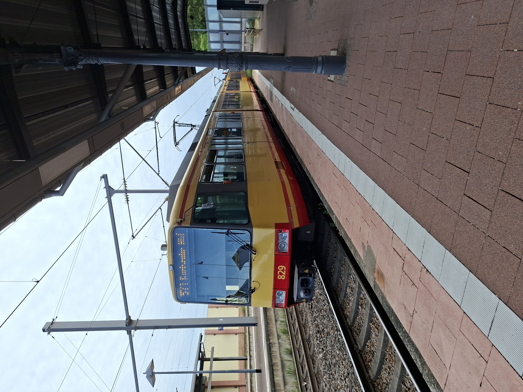 An exterior, eye-level, medium shot captures a yellow and red tram numbered 829 parked at a station in Sinzheim, Germany. The tram is positioned on the left side of the frame, facing right, with its front end in the foreground. The tram has a modern design with large windows and a prominent digital display on the front showing "571 Karlsbad Oststadt." The platform, paved with red brick tiles and accented by a white tactile paving strip, runs along the right side of the image. A dark grey, ornate metal pole supporting the station's roof stands prominently on the platform. The tracks are visible to the left of the tram, partially covered with gravel. Overhead, a complex network of catenary wires and support poles indicates the presence of an electric railway system. The sky above is overcast. In the background, a light-colored building with a sloped roof and a tall lamp post are visible. The scene suggests a quiet moment at a public transportation hub.