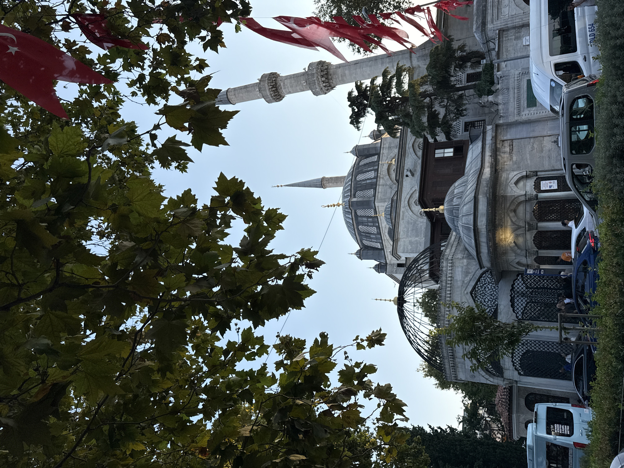 An exterior, eye-level shot shows the Mihrimah Sultan Mosque in Üsküdar, Turkey, from the west. The mosque complex is built of stone and features multiple domes and a tall, slender minaret topped with a conical spire. Red Turkish flags with white crescents and stars are strung across branches of trees in the foreground, partially obscuring the view of the mosque. The sky is clear and light blue. Vehicles, including a white van and several cars, are visible on a street in front of the mosque, and a few pedestrians are seen near the entrance. The foliage of large trees with broad leaves hangs over the top of the scene.