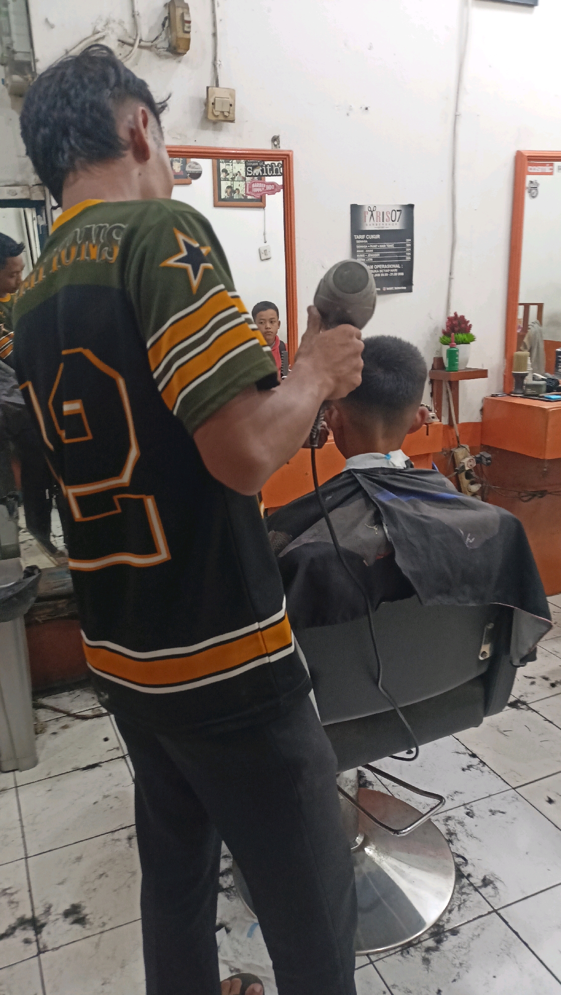 A barber is blow-drying the hair of a young male customer in a barbershop in Kertawangi, Indonesia. The barber, wearing a black and green jersey with orange and white stripes, holds a black hairdryer. The customer, seated in a gray barber chair and draped with a black cape, has a short haircut. In the background, a mirror reflects the scene, along with a framed picture and a price list poster. The floor is littered with cut hair.