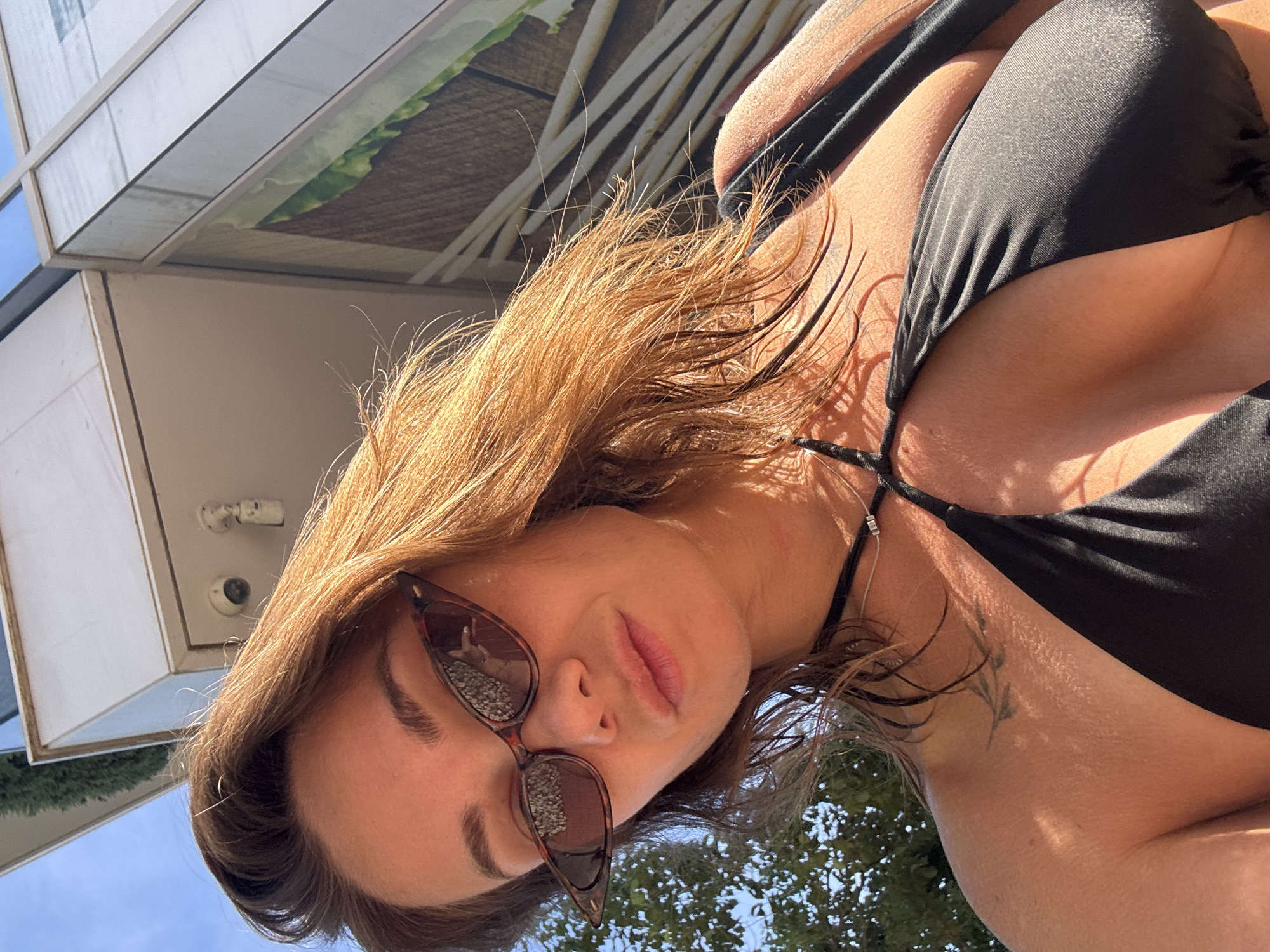 A woman in a black bikini is pictured from a low angle, looking up at the camera. She is wearing cat-eye sunglasses with a tortoise-shell pattern and a thin necklace. Her wet, reddish-brown hair frames her face. Behind her, a modern building with reflective windows and a textured wall can be seen against a bright blue sky. Sunlight casts shadows and highlights on her skin and hair. The scene appears to be outdoors, possibly in a city like Budva, Montenegro, given the architectural style and the sunny, urban environment suggested by the building.