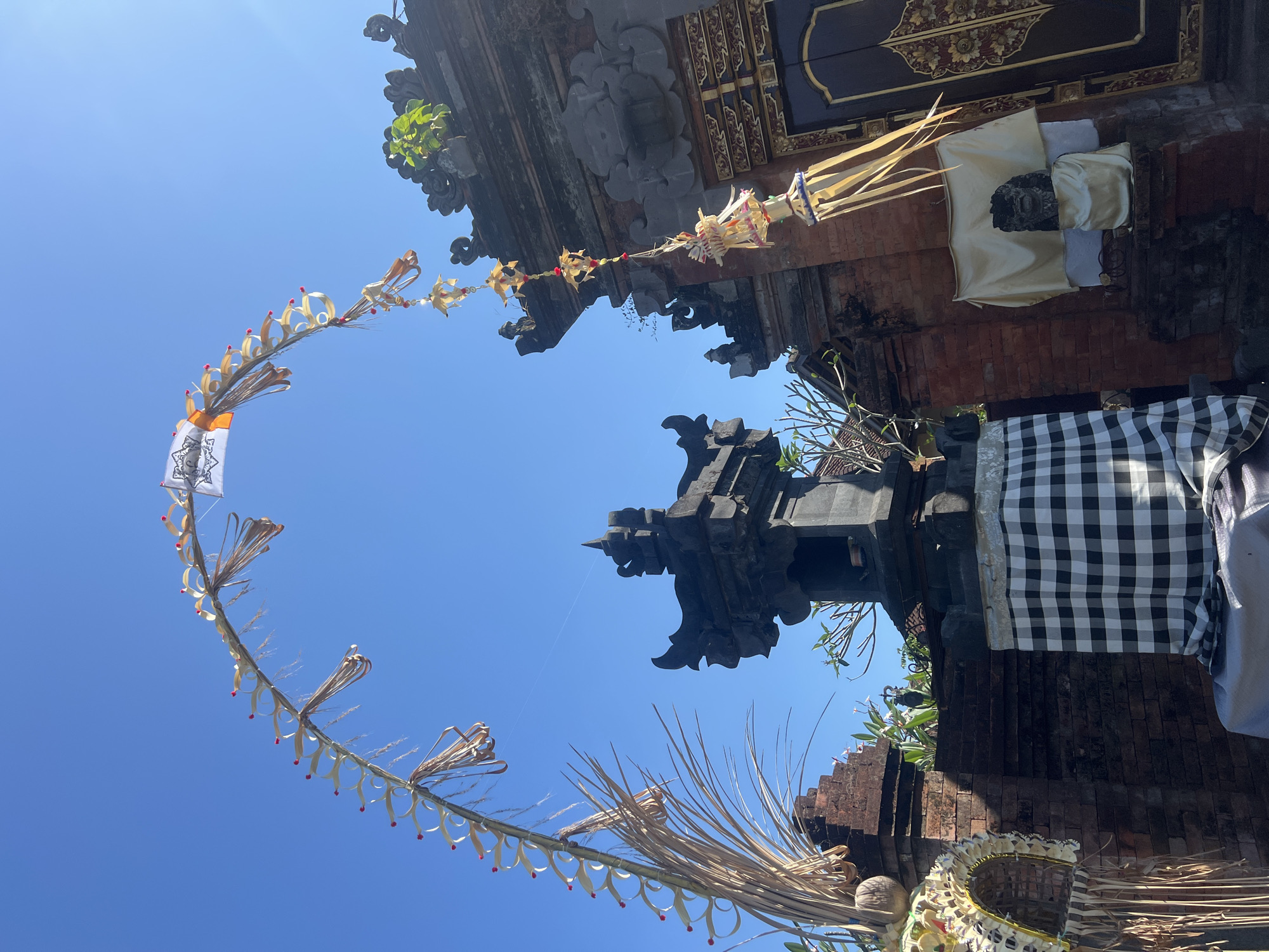 This media file depicts a vibrant outdoor scene at a temple in Sukawati, Indonesia, likely during daytime under clear, sunny skies. The primary subjects are the intricate decorations adorning the temple's architecture, characteristic of Balinese cultural and religious practices.
Dominating the frame are several ornate decorations. A long, crescent-shaped garland made of woven palm leaves and adorned with small red embellishments hangs prominently in the foreground, creating a welcoming arch. This garland is interspersed with white banners bearing intricate black patterns. Other woven decorations, possibly offerings or symbolic items, are visible near the temple structures.
The temple itself features traditional Balinese architecture with dark stone carvings and red brickwork. One section of the temple is draped with a large piece of fabric, a black and white checkered pattern commonly seen in Balinese temples. Another section reveals a stone statue, possibly a deity or a significant figure, covered with light yellow and white cloths. The overall setting suggests a place of worship or cultural significance.
The activity taking place is not explicitly shown, but the decorations strongly indicate a festive occasion or a daily ritual at the temple. The bright sunlight suggests it is likely midday. The clear blue sky contributes to a sense of peace and reverence associated with religious sites. The location cues, such as the architectural style and decorations, definitively place the scene in Sukawati, Indonesia, a region known for its rich cultural heritage and active temple life. No discernible text is visible in the image.