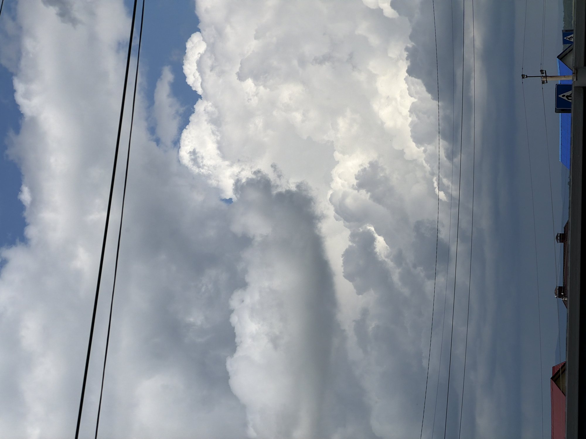 The image depicts a cloudy sky above a cityscape. Multiple black electrical wires stretch across the frame horizontally, obscuring parts of the sky. The sky is filled with a mix of white and gray clouds, with patches of blue visible. Below the wires, the roofs of several buildings are visible, some with red or blue roofing material. There are also traffic signs, one featuring a pedestrian crossing symbol, and what appears to be a light pole. The overall scene suggests an outdoor, urban environment under a dynamic sky.