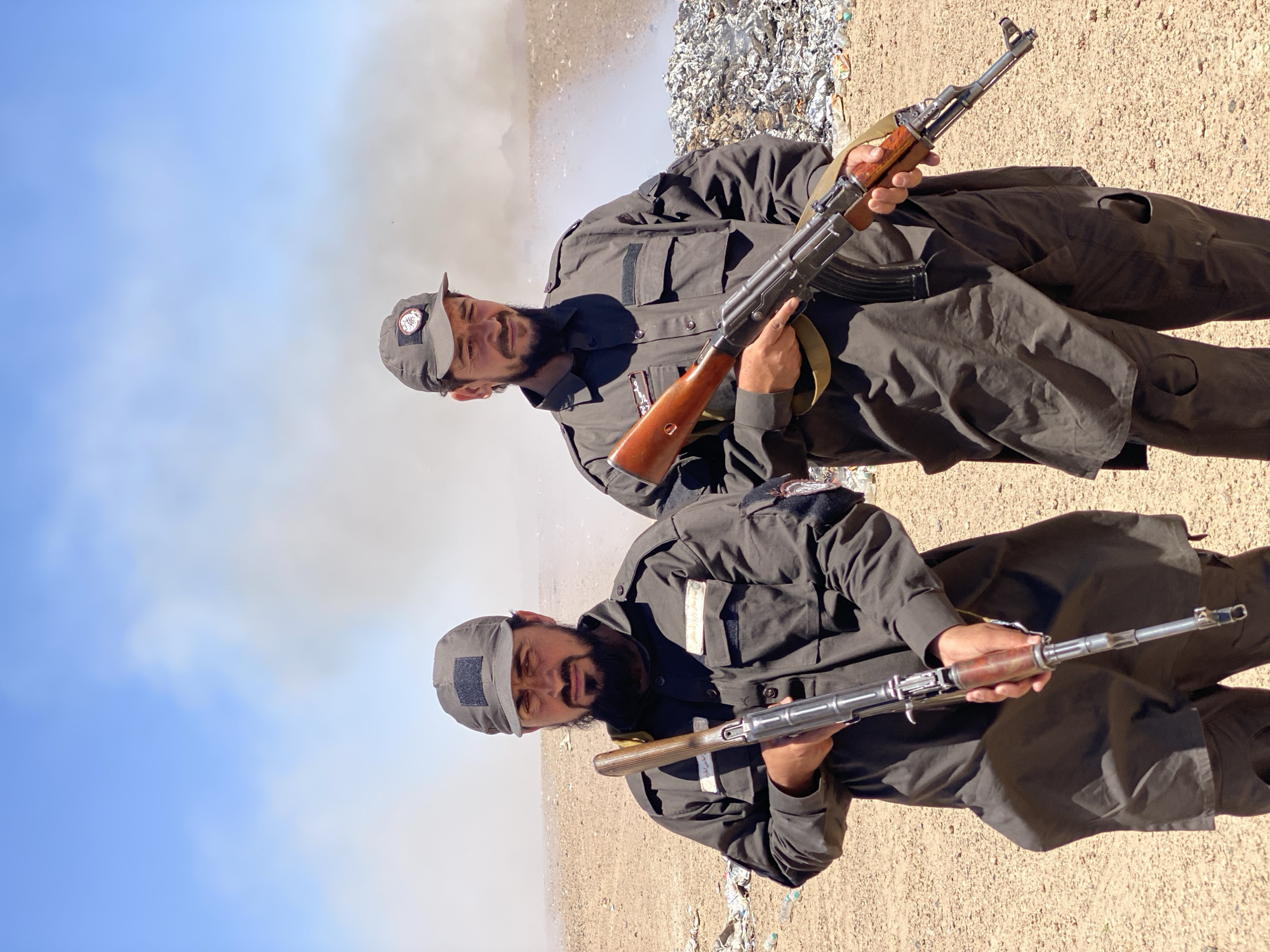 Two men in dark uniforms and caps stand in a barren, sandy landscape with a pile of debris and smoke in the background, indicative of a fire or explosion. The man on the left, with a beard, holds an older model rifle with a wooden stock. The man on the right, also bearded and wearing a cap with a circular emblem, holds an AK-47 style rifle. The environment appears to be outdoors, possibly a remote area or a landfill site. The sky is blue with some haze. The uniforms are dark, possibly military or security forces, with patches on the sleeves and caps. The man on the left has text on his uniform above his left pocket. The man on the right has a circular patch on his cap with what appears to be an emblem. The presence of firearms suggests a security or military context. The debris in the background might be trash or wreckage. The smoke could be from a controlled burn or an uncontrolled fire. The overall impression is a scene of some activity in a rugged environment.
