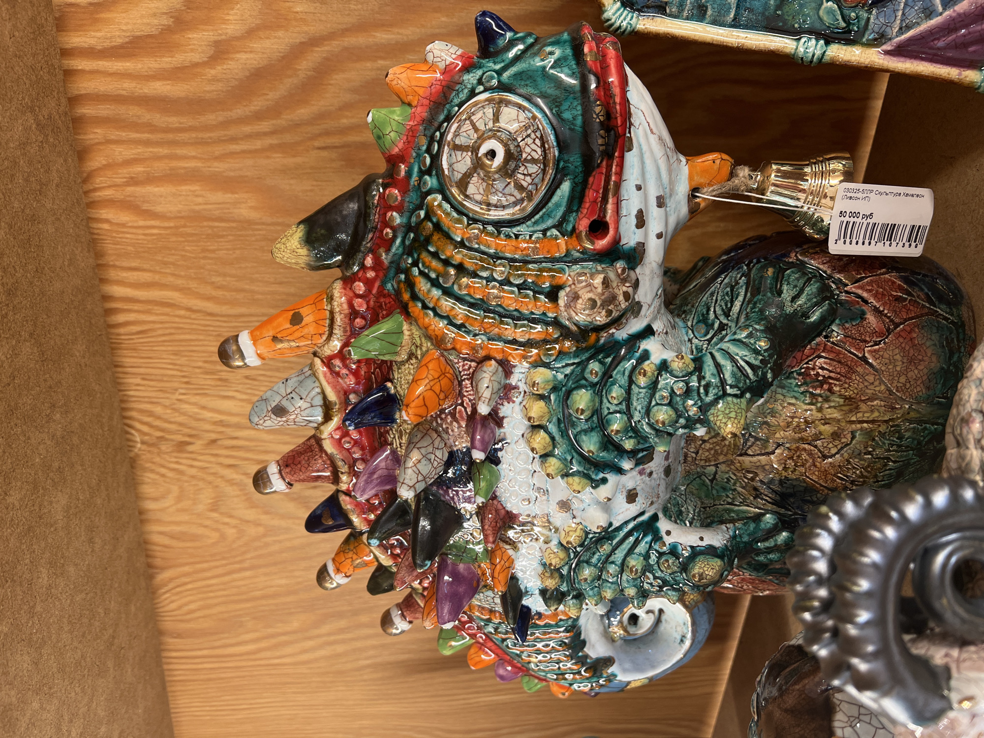 The image shows a highly decorated ceramic sculpture of a chameleon. The sculpture is colorful with a variety of textures and patterns and glazed with orange, green, blue, and white. It appears to be a handcrafted item with intricate details. A price tag is attached to the chameleon, indicating that it is for sale. It says 50 000 rub. The item is located in Moscow, Russia. The background consists of a light wood-colored surface.