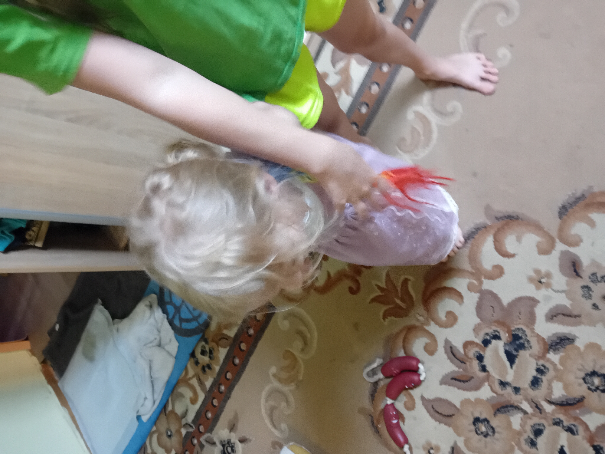A toddler with blonde hair is wearing a pink dress and is being held by a person wearing a green shirt and yellow shorts. The toddler is reaching for an orange toy. They are standing on a patterned carpet in what appears to be a room. To the left of the toddler, there are red toy shoes. In the background, a blue and white object is visible, possibly a mat. A dark gray garment is also visible behind the blue and white object. The scene suggests an indoor setting in Chuhuiv, Ukraine.