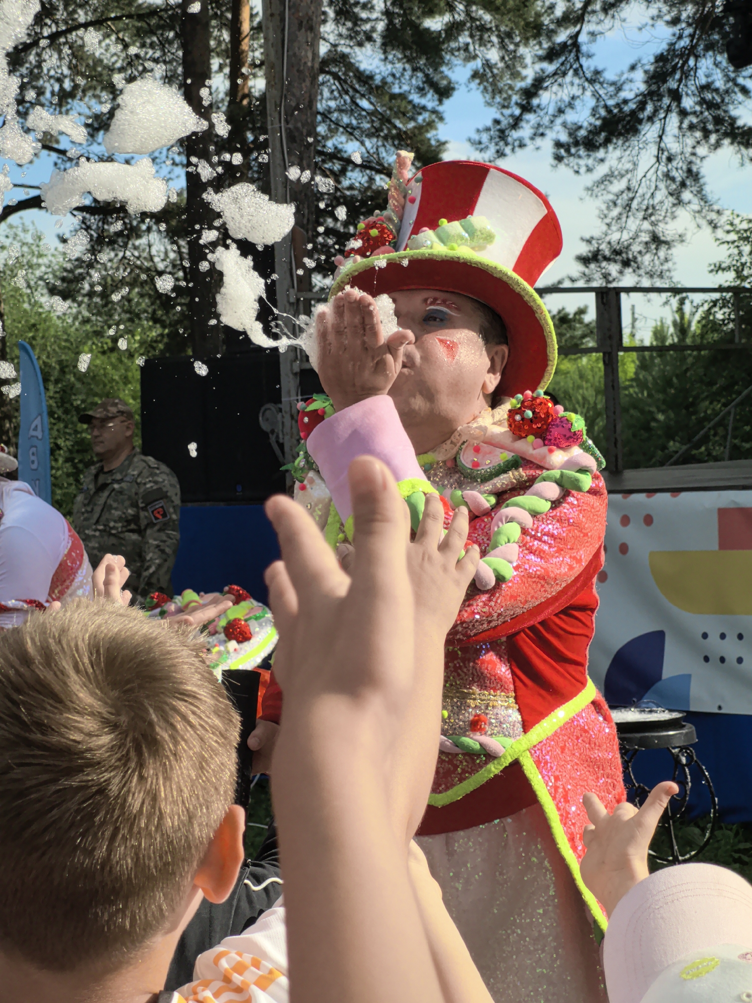 In the city of Gorniy, Russia, a performer in a vibrant, sequined red and yellow costume with a tall, striped hat adorned with colorful decorations blows a stream of bubbles toward the viewer. Their face is lightly made up with glitter and red markings. In the foreground, the hands of children are reaching out towards the bubbles. Behind the performer, a man in a camouflage military uniform stands attentively. The scene takes place outdoors, with trees and a clear blue sky visible in the background, suggesting a sunny day and an outdoor event or performance.