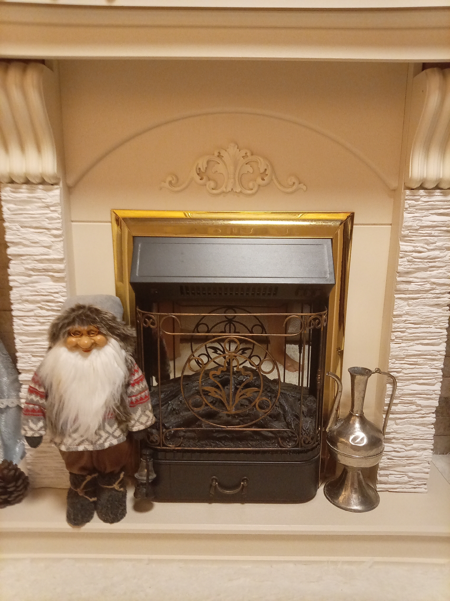 A festive Christmas gnome decoration sits to the left of an electric fireplace. The gnome is wearing a grey hat, a red and grey patterned sweater, and brown pants with dark grey boots. The fireplace is framed in gold and has a black grate with decorative metalwork. To the right of the fireplace is a silver pitcher. The fireplace and decorations are placed on a mantlepiece. This image was taken in Yessentuki, Russia.