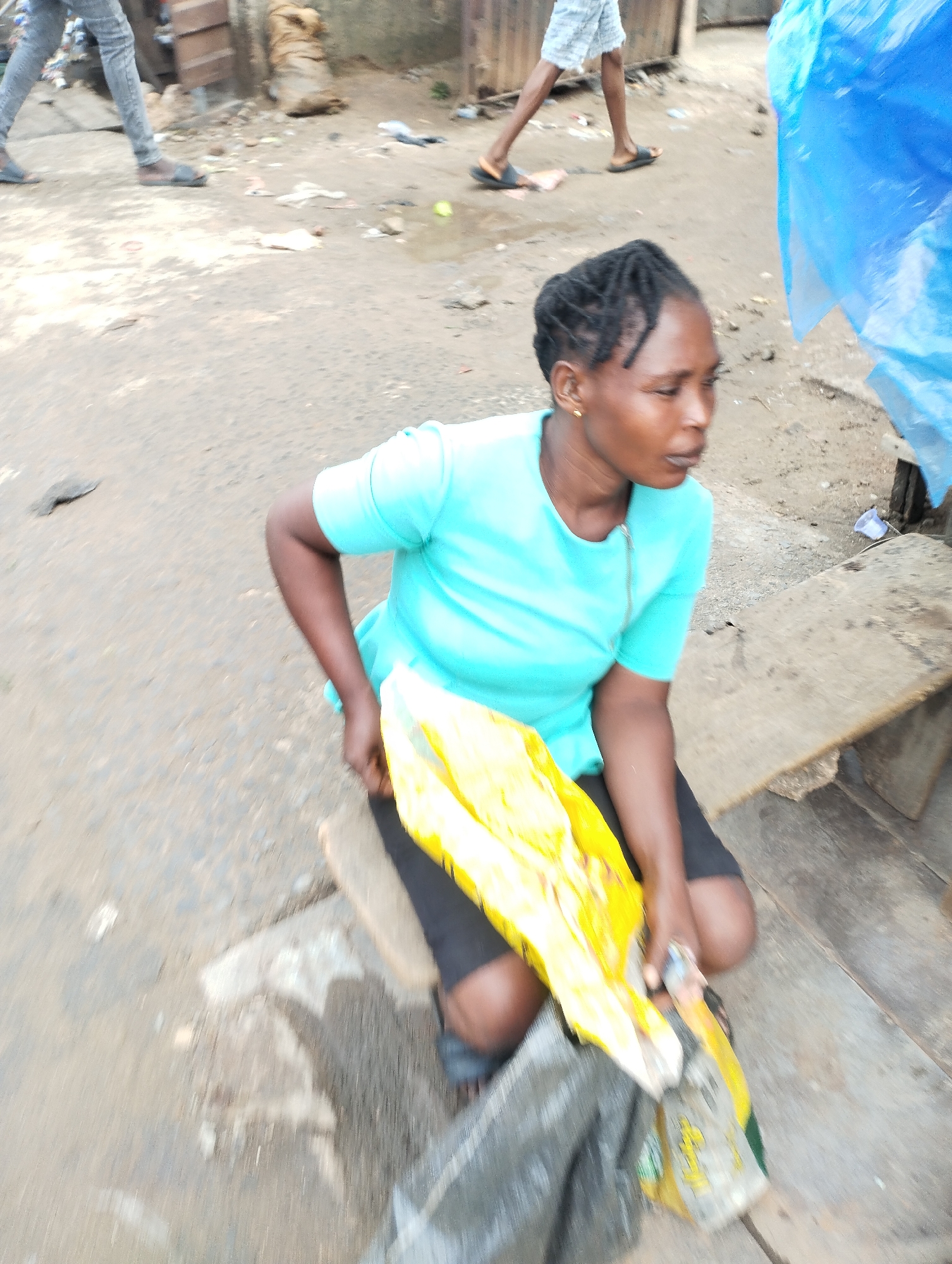 In Fegge, Nigeria, a woman with braided hair and wearing a light blue shirt and black shorts is kneeling on a low stool. She is holding a yellow plastic bag, partially obscured by a black bag. The ground is paved and littered with debris. Two people are walking in the background. A piece of blue plastic hangs from a structure in the background. Wooden structures are also visible in the background.