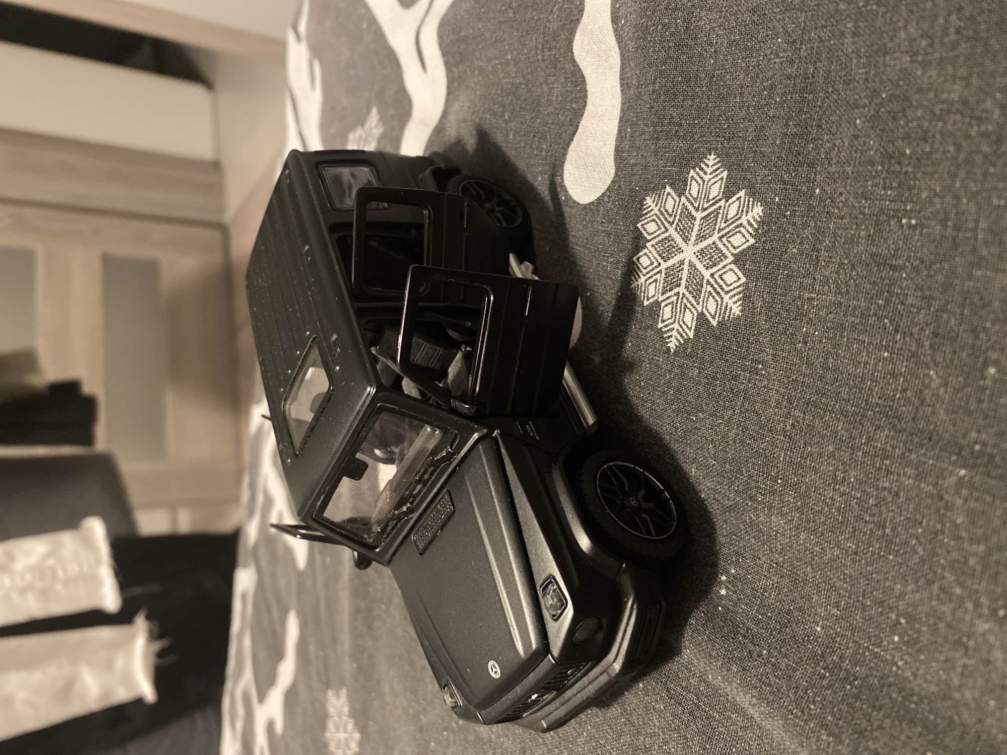 A close-up, eye-level, indoor, full shot shows a black, die-cast model Mercedes-Benz G-Wagon with its doors open, placed on a dark grey fabric with a white snowflake pattern. The model car, oriented diagonally from the lower left to the upper right, is the primary subject. Its hood, front grille with the Mercedes-Benz logo, headlights, side mirrors, open driver and passenger doors, and visible interior details such as seats and steering wheel are clearly depicted. The car rests on a patterned surface that appears to be a blanket or duvet. In the blurred background, there are hints of furniture and possibly clothing hanging, suggesting a domestic setting. The lighting is somewhat dim, casting soft shadows. The specific location in Łódź, Poland, cannot be determined from the provided image.