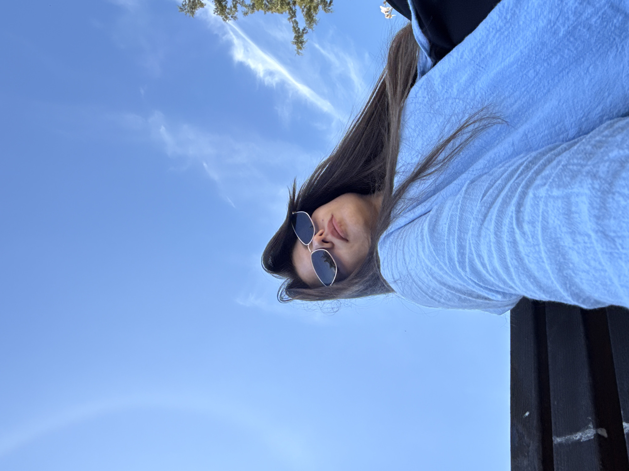 A low-angle, close-up shot captures a young woman from the chest up, sitting on a dark wooden bench under a clear blue sky with scattered wispy clouds. The woman has long, dark brown hair that flows over her shoulders and is partially obscuring her face. She is wearing cat-eye sunglasses with reflective lenses and a light blue, textured long-sleeved top. The background is dominated by the sky and a hint of green foliage from a tree on the right side of the frame. The lighting suggests it is daytime, likely sunny, as indicated by the bright sky and the reflections on her sunglasses. The overall impression is casual and relaxed, possibly taken outdoors during the day.