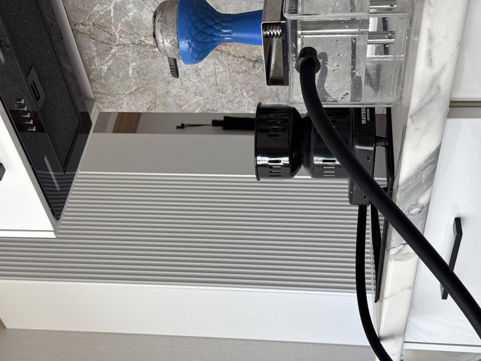 The scene captures a detailed view of a modern hookah setup situated on a white marble countertop with grey veining, in what appears to be a kitchen or recreational area in the city of Sochi, Russia. A transparent, square-based hookah with water and internal metal tubing is present on the right, connected by a black silicone hose that extends across the countertop. A bright blue, textured hookah bowl, topped with a metallic heat management device containing charcoal, rests atop the hookah. To the left of the hookah, a black electric charcoal burner, branded "WAYSKO ceramics," sits on the countertop. The background features a greyish-brown mottled stone or tile backsplash. Above, a black range hood with a control panel, displaying several red indicator lights and silver knobs, is mounted. White base cabinets with black rectangular handles are visible below the countertop. To the far left, a white wall with distinct vertical fluted paneling is present. The overall lighting is bright and even, indicating an indoor setting.