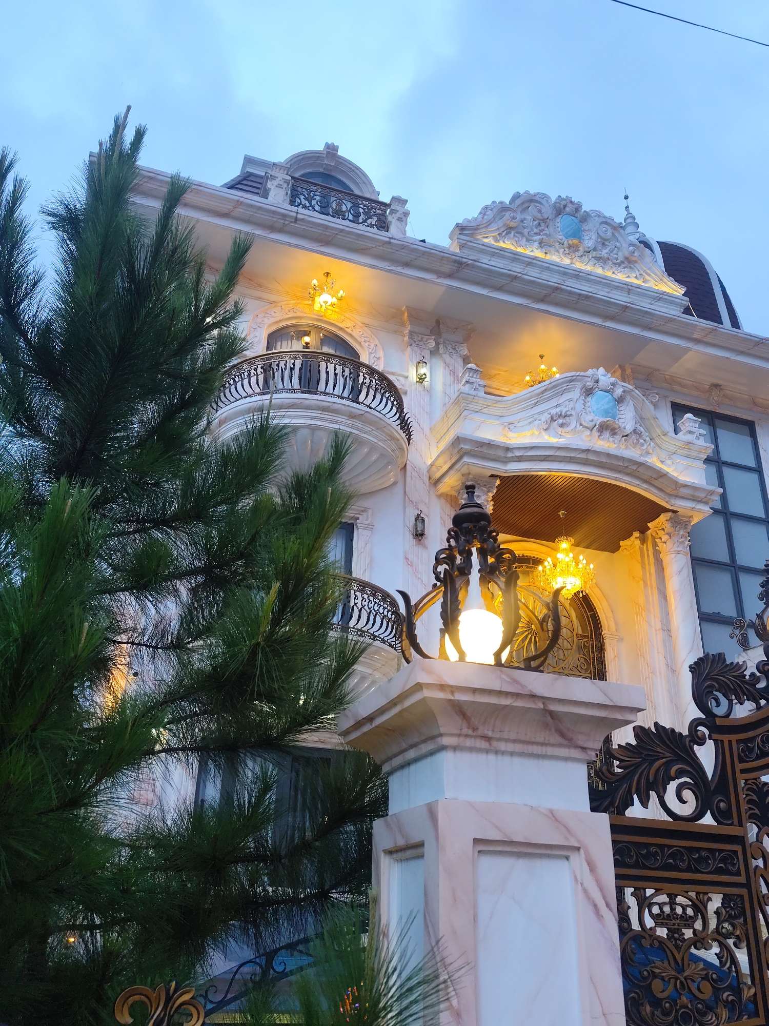 A low-angle, close-up outdoor shot shows the facade of a luxurious, ornate building with intricate architectural details and a dark sky overhead. The building's exterior is adorned with white marble, accented with pink veining, and features multiple balconies with black wrought-iron railings. Ornate decorative carvings, illuminated by warm yellow lights, are visible along the roofline and around arched windows. A large, detailed chandelier hangs under an overhang. In the foreground, a pine tree with long needles partially obscures the view of the building. To the right, a black wrought-iron gate with elaborate scrollwork and crown motifs is visible. The scene is set in the city of Xuân Trường - Đà Lạt, Vietnam. There are no people or animals present in the image.