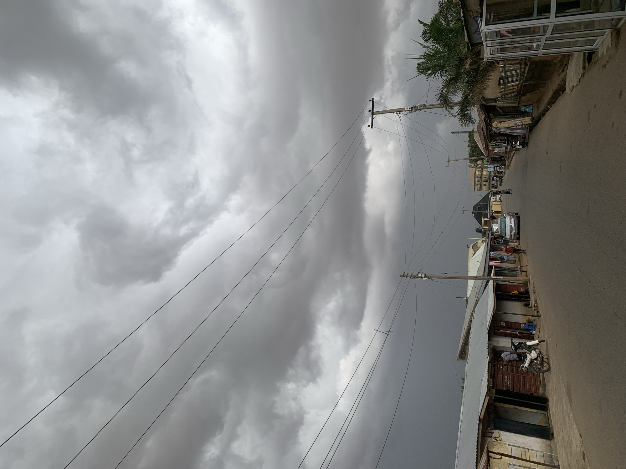 The image presents a street view in Jos, Nigeria, dominated by a heavily overcast and ominous sky. The upper two-thirds of the frame are filled with dense, dark grey clouds, swirling and layered, suggesting an impending storm or persistent gloomy weather.
Stretching across the sky are numerous utility poles and a complex network of power lines, which run diagonally from the right side of the frame towards the left, contrasting starkly against the monochrome clouds. Along the right side of the image, a paved road extends into the distance, bordered by a continuous line of buildings. These structures, many with corrugated metal roofs and weathered facades, appear to be a mix of residential and commercial establishments, with some storefronts visible. Palm trees add a touch of greenery near the top right corner.
On the street, a white car is visible further down the road, and a white motorcycle is parked nearer to the foreground, close to one of the buildings. A few indistinct human figures can be seen walking or standing on the street or near the buildings, but their details are not clear. The overall lighting is dim, indicative of a very cloudy day, possibly late afternoon or an early morning overshadowed by thick cloud cover. The scene evokes a quiet, somewhat somber atmosphere due to the heavy skies. No discernible text is visible on any signs or structures.