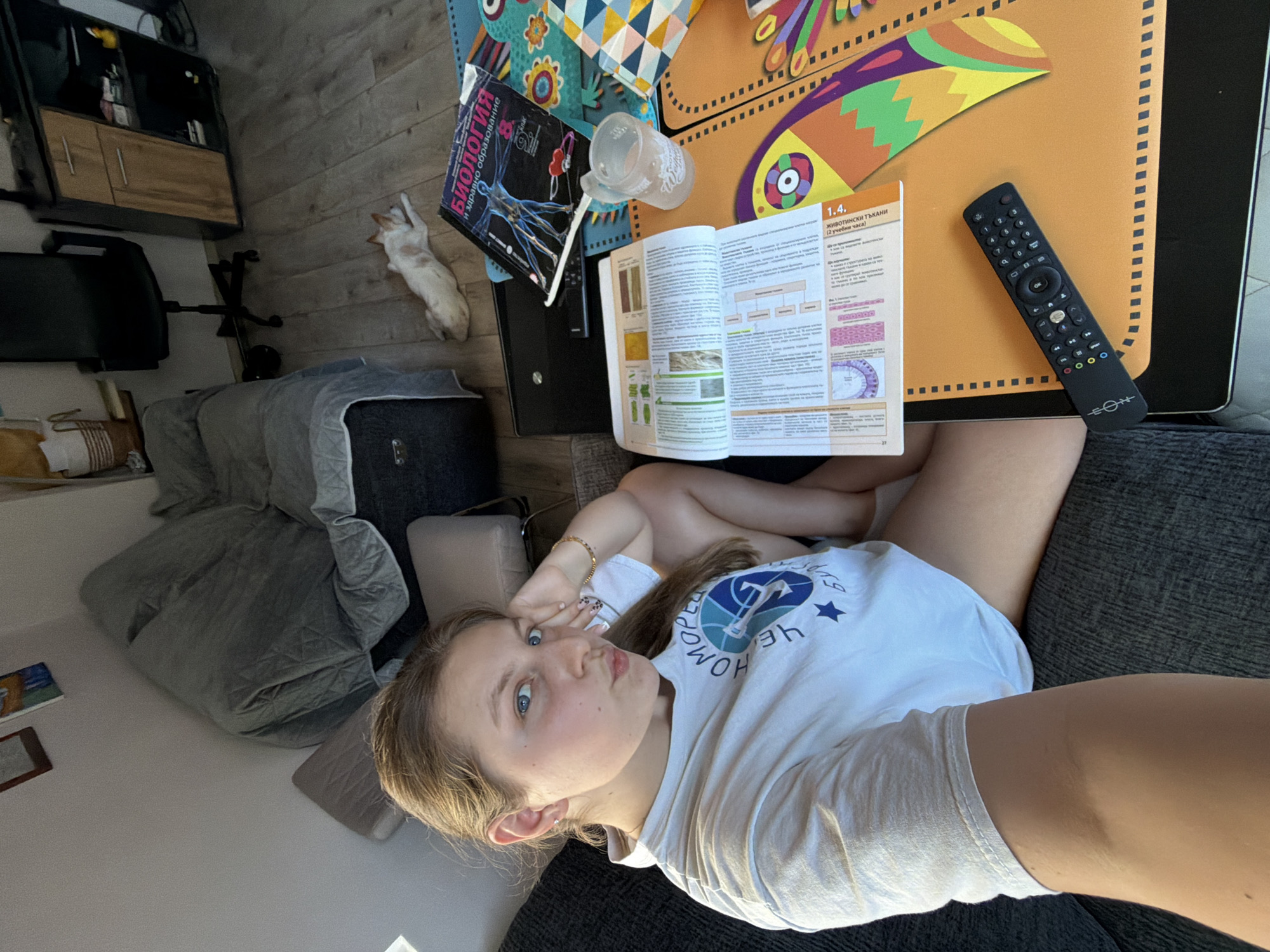 The image depicts an indoor scene, likely a living room or study area. A young woman with light brown hair, wearing a white t-shirt, is seated on a dark gray sofa. She is looking at the camera with her left hand resting on her head and her right arm bent. She is pouting, with her lips pursed. Beside her, on a coffee table, are several open books, a glass mug, and a television remote control. A cat is lying on the floor in the background. The room appears to be cluttered with furniture, including a desk with a chair, and a covered sofa or armchair. There are framed pictures on the wall. The presence of a cat, books, and a remote control suggests a casual, domestic environment. The style of the furniture and decor does not provide any specific clues to identify the city of Burgas, Bulgaria, or any other location.