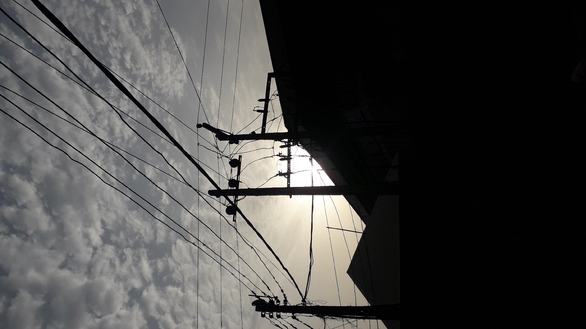 The image shows a low-angle view of electricity poles and wires silhouetted against a cloudy sky. A building's dark silhouette occupies the right side of the frame, obscuring much of the background. The sun appears to be shining through a gap in the building, creating a bright spot. The image was taken in a location with power lines and infrastructure, possibly within a city. I cannot verify that the image was taken in Onitsha, Nigeria.