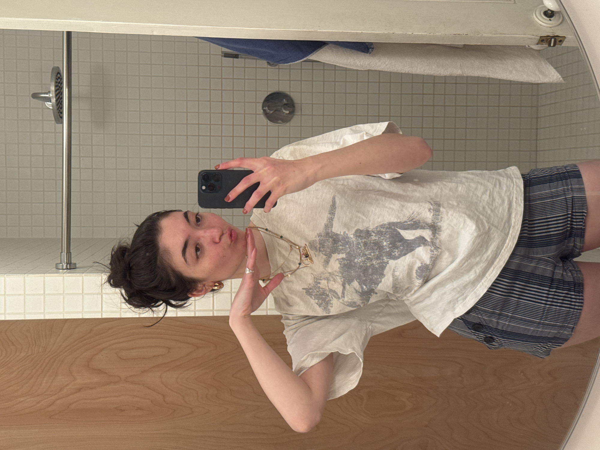 A young woman is captured in a mirror selfie, posing playfully in what appears to be a bathroom setting. The image is oriented vertically, with her head towards the top. She has dark hair pulled back into a messy bun and is making a "fish face" with pursed lips, looking directly into the camera. Her left hand is raised to her chin in a relaxed gesture, while her right hand holds a dark-colored smartphone, likely an iPhone, to take the picture.
She is wearing a light-colored (possibly off-white) t-shirt featuring a dark grey graphic print, which appears to depict an animal like a horse. Her attire also includes blue and white striped shorts with visible buttons on the side. She accessorizes with a gold-toned necklace that has small beads and a distinctive clasp, and gold hoop earrings. Her fingernails are painted with light brown polish.
The background, visible through the mirror's reflection, reveals a tiled wall covered in small white square tiles, indicative of a shower or bathroom area. A silver showerhead and faucet controls are visible on this wall. Above the tiled section, a blue towel and a white towel are draped, and part of a white door with a round doorknob can be seen. A warm-toned wooden counter or vanity surface occupies the foreground of the reflection. The overall impression is casual and cheerful. No visible text is legible in the image.