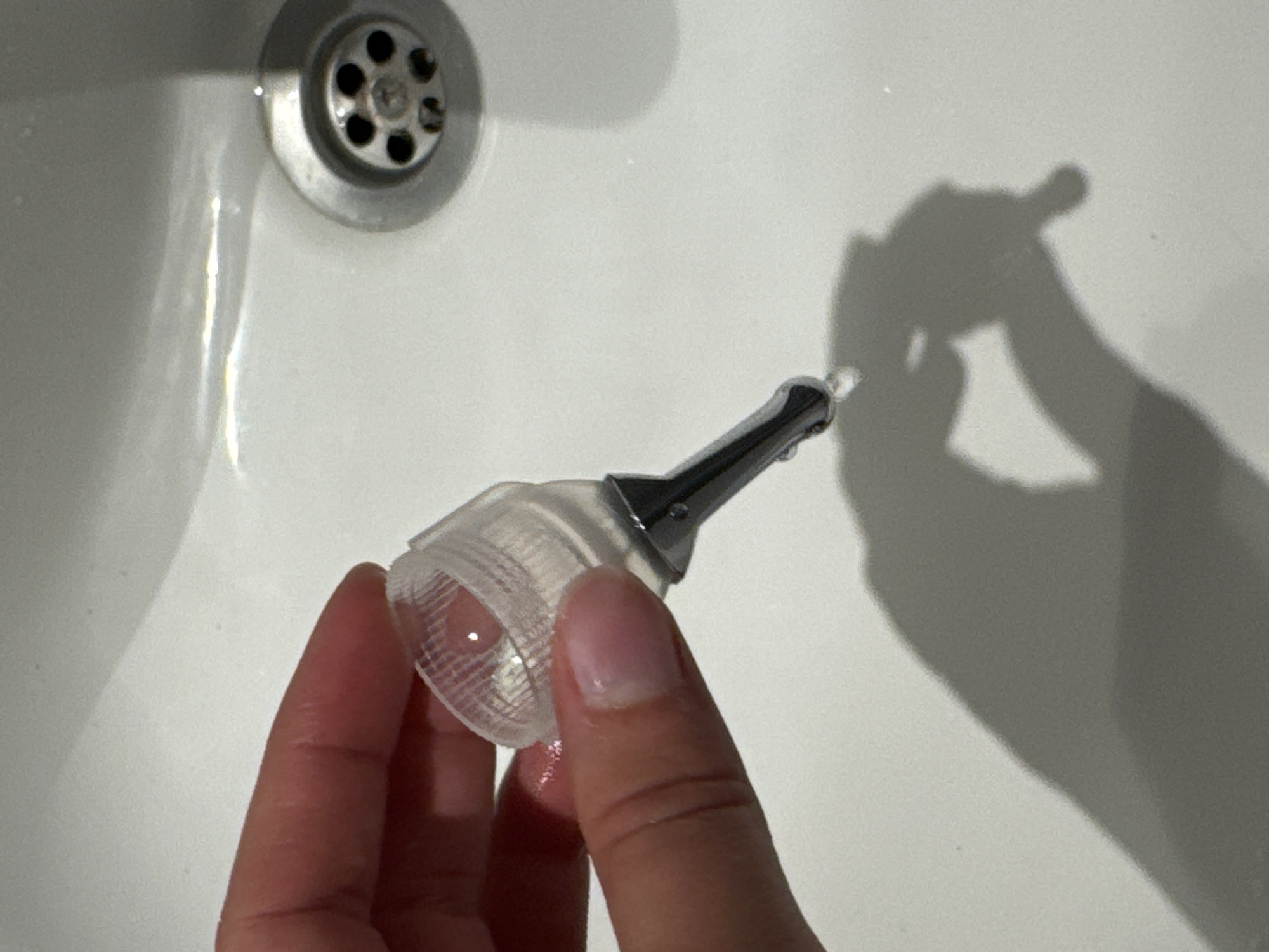 A close-up shot shows a person's hand holding a transparent, ridged cup with a metal nozzle attached. A small stream of water is dripping from the nozzle onto a white sink. The sink has a metal drain with a circular grate visible to the right of the object. The lighting creates a distinct shadow of the hand and object on the sink's surface. The image does not provide any specific location within Aksu, Turkey.