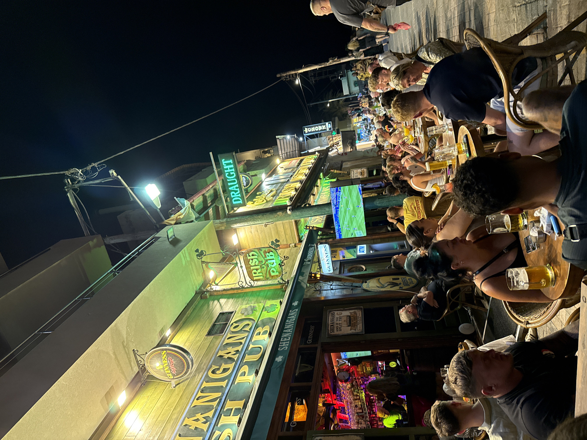 The image depicts an outdoor dining area at "Shenanigans Irish Pub" in Chersonisos Municipal Unit, Greece, at night. The pub is brightly lit with green and yellow neon signs advertising "Shenanigans Irish Pub" and "Draught". People are seated at tables on a paved walkway, many of them consuming alcoholic beverages, primarily beer. Some patrons are engaged in conversation, while others appear to be watching a television screen displaying a soccer match. The atmosphere is lively and social, suggesting a popular establishment. The architecture of the buildings is typical of a tourist-oriented coastal town, with multiple stories and balconies. The lighting is a mix of artificial sources, casting a warm glow on the scene.