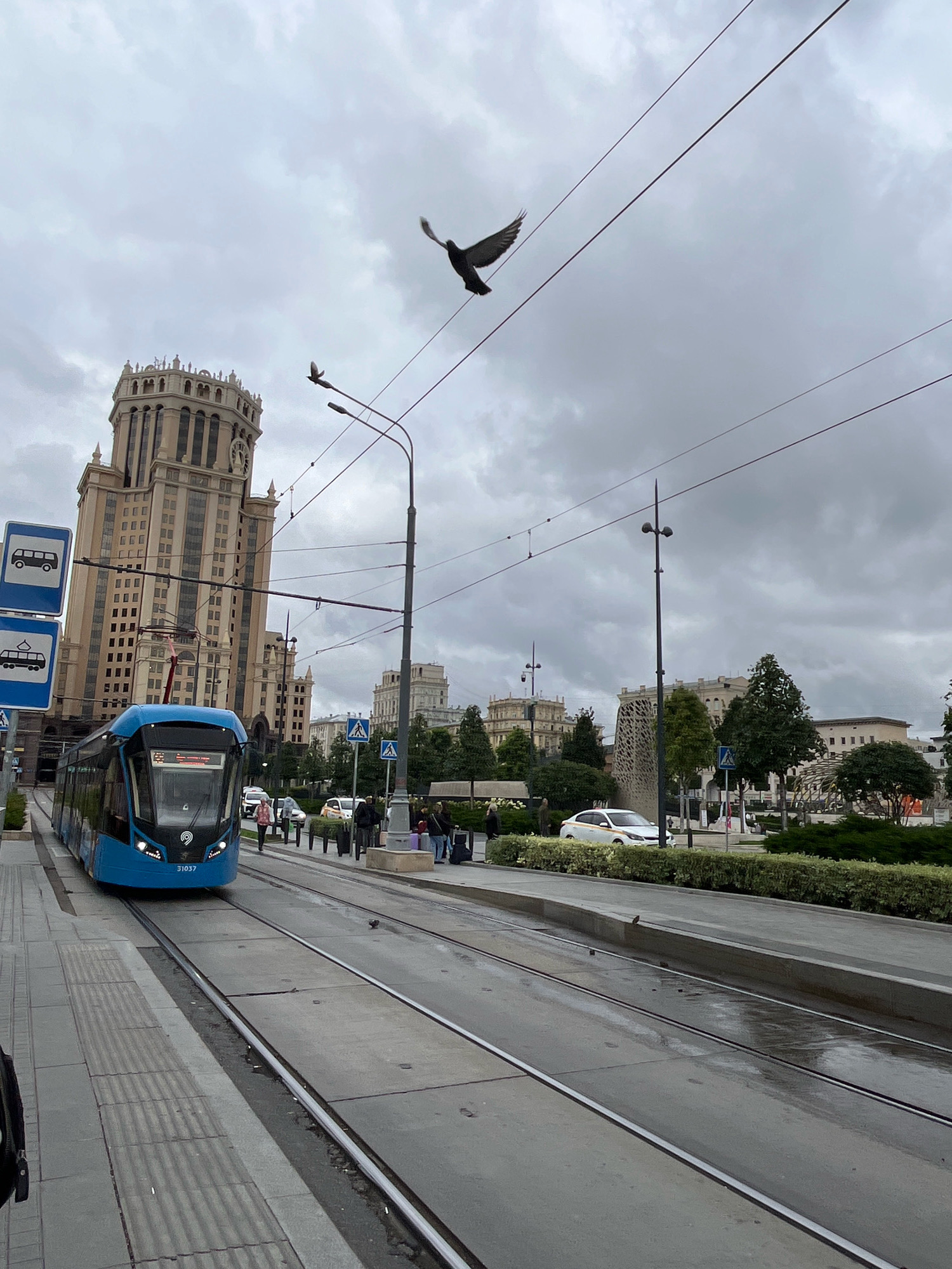 The image captures a street scene in Moscow, Russia, dominated by a distinctive Stalinist skyscraper. In the foreground, a modern blue tram with the number 31037 is positioned on tram tracks. Several people are visible near the tram stop, some with luggage, suggesting they are waiting for or have just disembarked from transportation.
A prominent traffic sign with bus icons is mounted on a pole to the left of the tram. In the background, the aforementioned skyscraper, characterized by its ornate architecture and clock tower, stands tall against a cloudy sky. Other buildings, trees, and decorative structures are also present, creating an urban landscape.
Two birds are in flight above the tram tracks and overhead wires. The ground appears wet in places, possibly from recent rain. The overall atmosphere suggests a typical day in a bustling city environment.