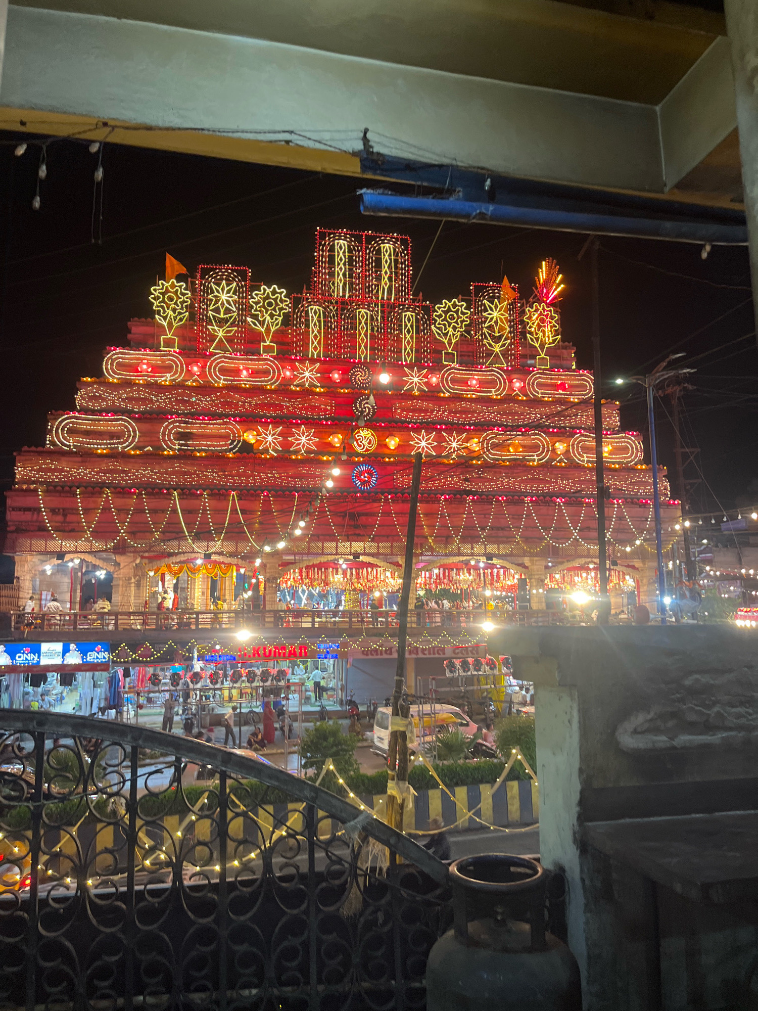 This is a night photograph of a tiered, elaborately decorated structure, likely a temple or a festive display, illuminated with numerous red and yellow lights. The structure has a layered, pyramid-like shape with intricate patterns of lights forming borders and decorative motifs, including starbursts and what appear to be stylized flowers. Yellow flags and tall, flame-like light fixtures are visible at the top.
Below the main structure, there are multiple levels of awnings and open spaces, with people visible on balconies and walkways, suggesting it's a public space with shops or stalls. Signs with text, some in English and some in Hindi, are partially visible. One sign clearly reads "KUMAR".
In the foreground, a black wrought-iron fence with decorative scrollwork is visible, with strings of warm yellow lights draped over it. Beyond the fence, there is a street scene with a white van, a rickshaw, and people walking. A dark gray gas cylinder is prominently positioned in the lower right corner of the frame, in front of a concrete structure.
The overall impression is of a vibrant, festive nighttime scene, likely during a cultural or religious celebration in Indore, India. The style of decoration, including the tiered structure and the lighting, is characteristic of festivals like Diwali or similar religious events held in India.