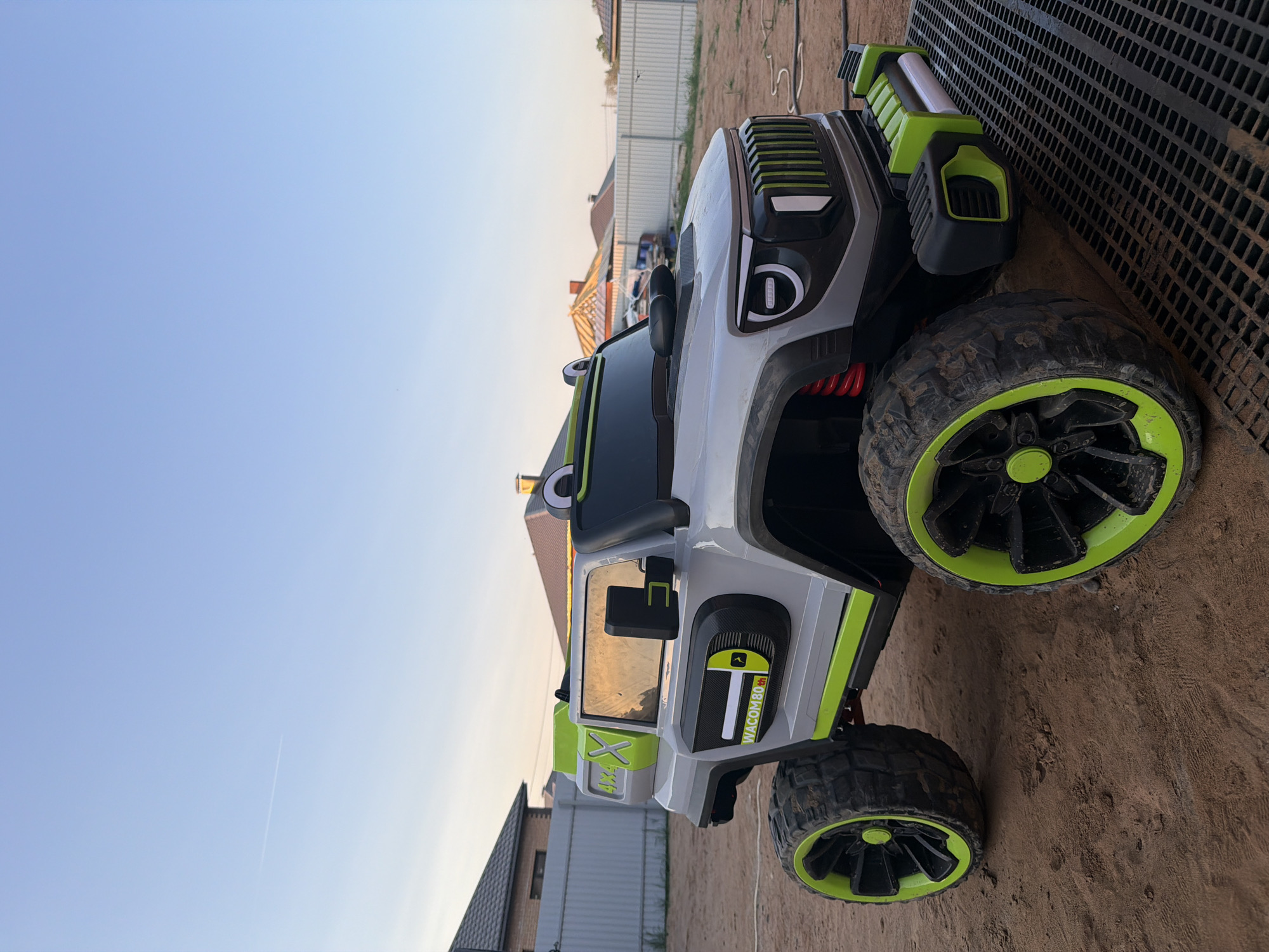 A slightly tilted, medium shot depicts a toy, off-road vehicle positioned outdoors. The vehicle, primarily gray with green accents, features large, black tires with green rims. It is situated on a surface covered with dark dirt, near a black grate. In the background, there are various houses, trees, and fencing under a clear blue sky. The photo was taken in Chaikovsky, Russia.