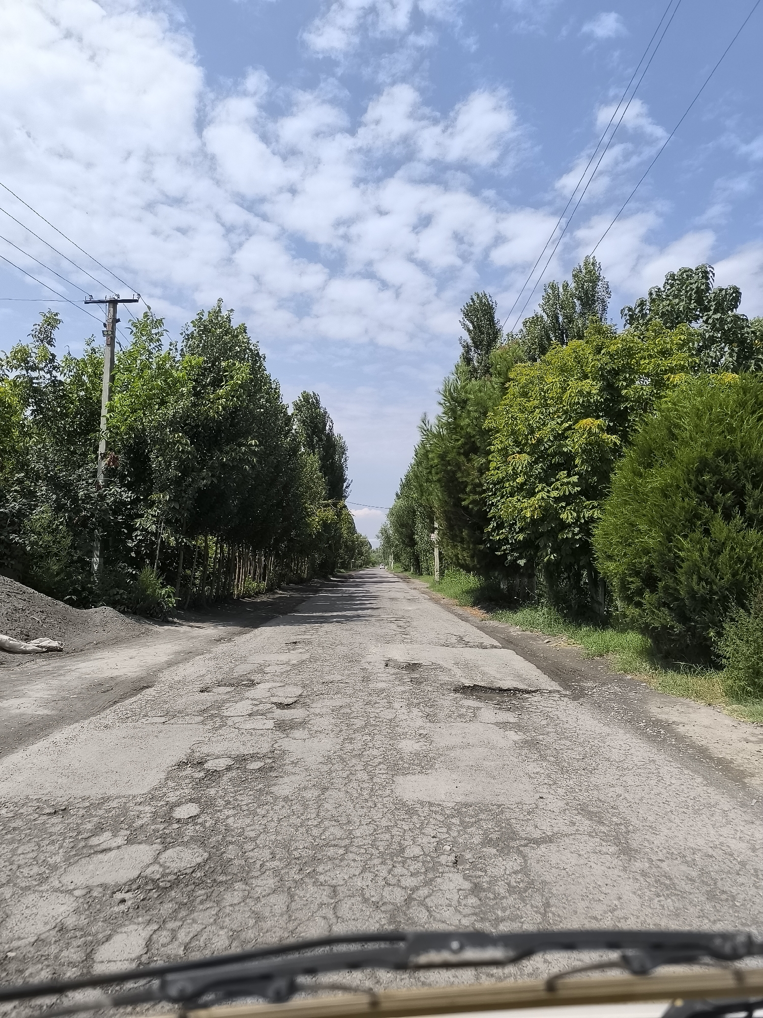 The image shows a rural road in the city of Shabada, Uzbekistan. The road is cracked and has many potholes. It is lined with trees and vegetation on both sides. There are power lines and a utility pole visible on the left side of the road. The sky is blue with scattered white clouds. The image appears to have been taken from inside a vehicle, as the windshield wipers and part of the dashboard are visible in the foreground.