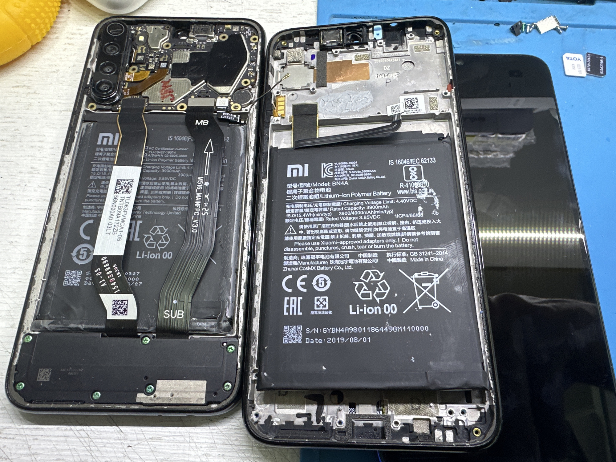 This media file is a close-up, top-down photograph of the interior of a disassembled smartphone, likely a Xiaomi device given the visible branding and text. The subjects are two separate phone components: the main logic board and the battery. The scene is a workshop or repair setting, indicated by the blue surface beneath the components, which appears to be a workbench, and some small electronic parts scattered nearby, including what looks like two SIM cards.
The activity taking place is a phone repair or disassembly. The components are laid out, revealing intricate circuitry and the large lithium-ion battery. One of the phone's main flex cables is labeled "MB," and another component is marked "SUB." The battery itself has extensive text printed on it, including its model number (BN4A), capacity (3900mAh), voltage (3.85VDC), manufacturer information (Zhuhai CosMX Battery Co., Ltd.), and a manufacturing date of "2019/08/01". There are also warnings in multiple languages about proper usage and disposal of the battery. Another flex cable connected to the logic board is labeled "M516_MAINFPC_V3.0". A partially visible second phone, entirely black, lies to the right, suggesting it is the original casing or another phone being worked on.
There are no people visible in the image. The lighting appears to be artificial, likely from overhead shop lights, and there are no indications of the time of day or weather. The location context of Moscow, Russia, is not directly evident from the image itself but is provided as external information.