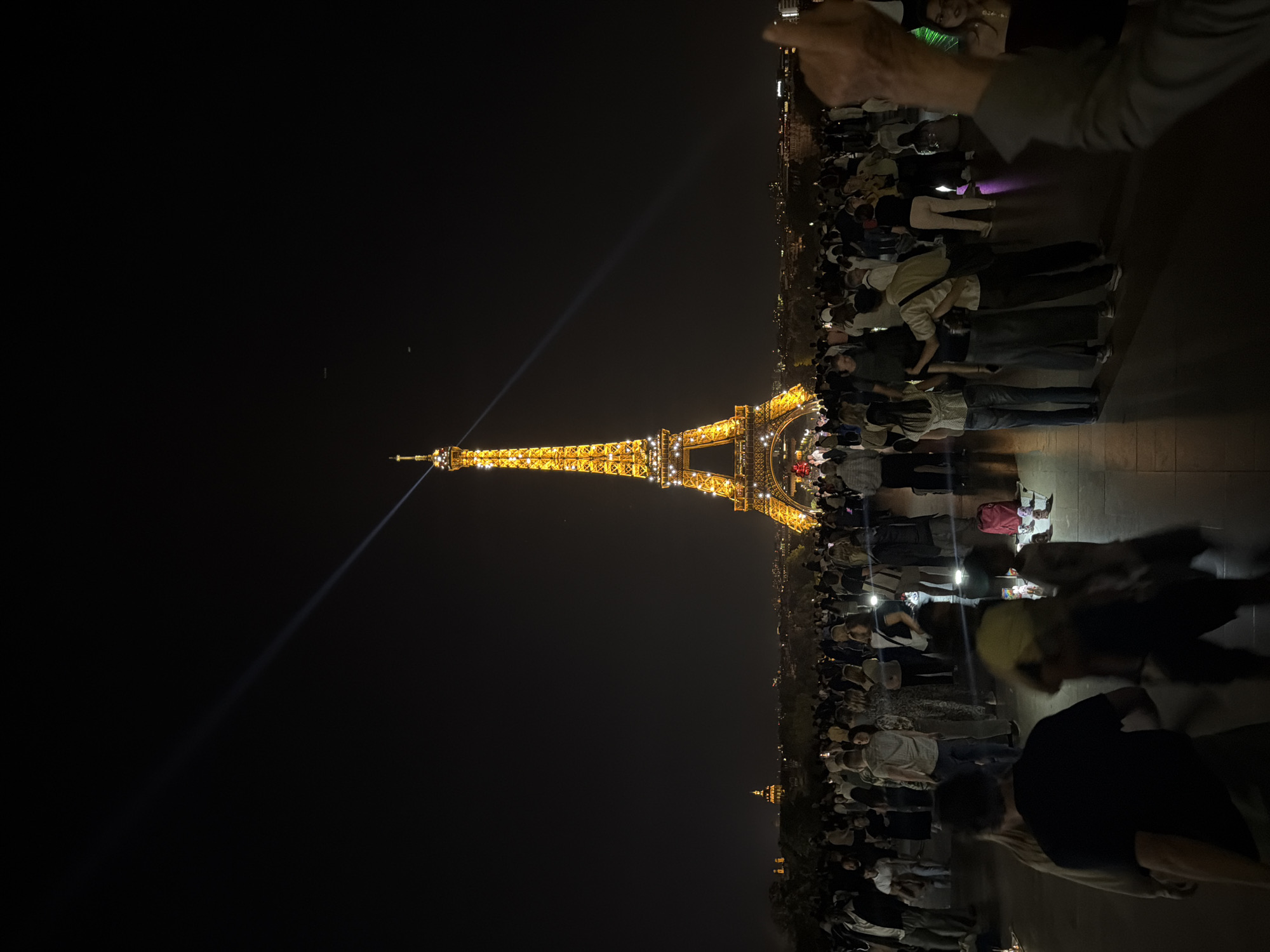 At night, the Eiffel Tower is brightly lit with golden lights, casting a beam of light into the night sky. A crowd of people stand in front of the tower, their faces illuminated by the tower's glow. The scene is taken in Paris, France.