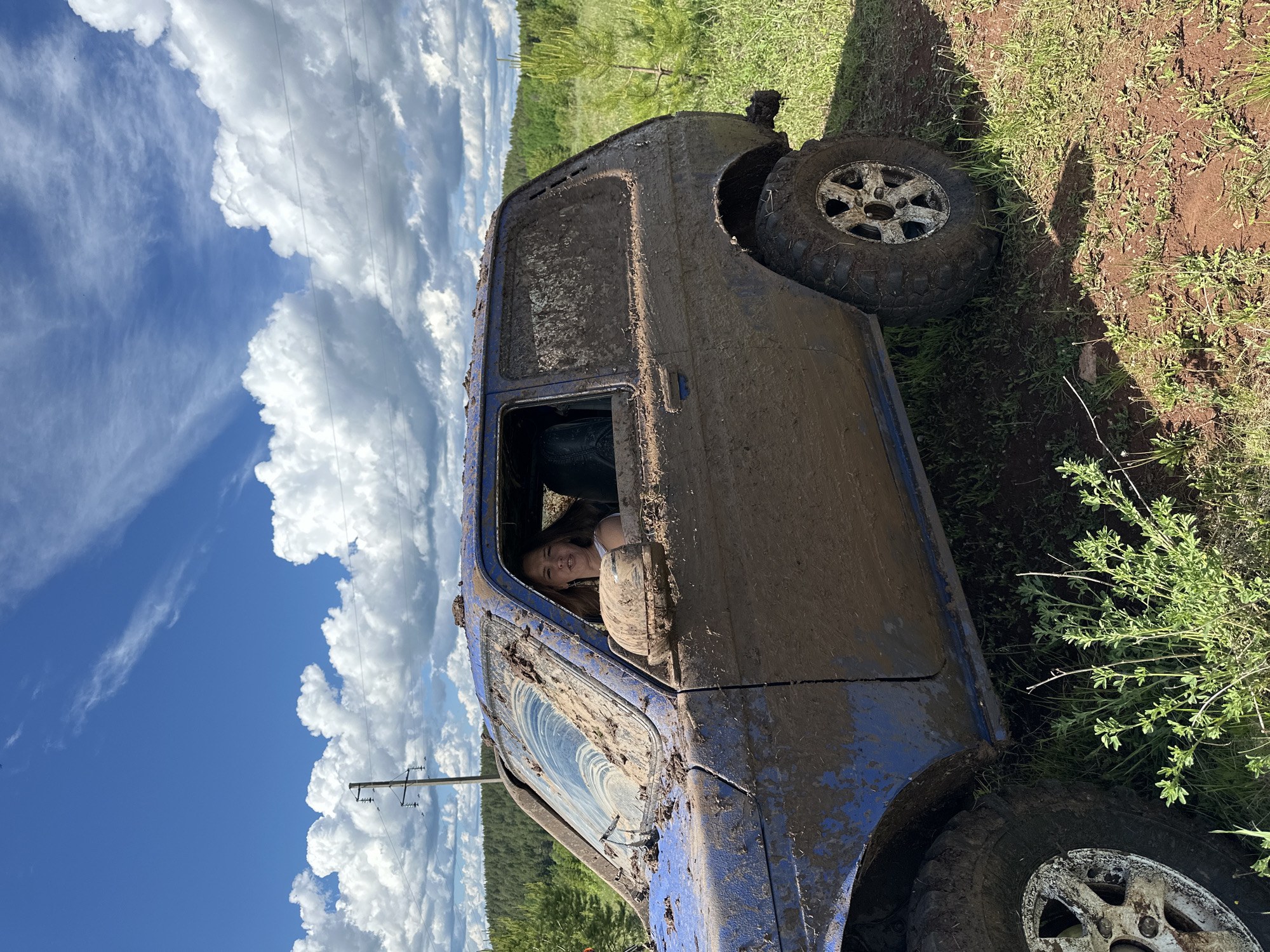 A blue vehicle, heavily coated in mud, is situated off-road in a grassy, dirt environment. A person is visible inside the vehicle, looking out of the driver's side window. A cloudy sky is in the background along with a power line in the distance. The ground to the right of the vehicle appears to be a grassy area with patches of dirt. The location of the image is consistent with a rural, off-road area, possibly near the town of Ilyinsky, Russia.