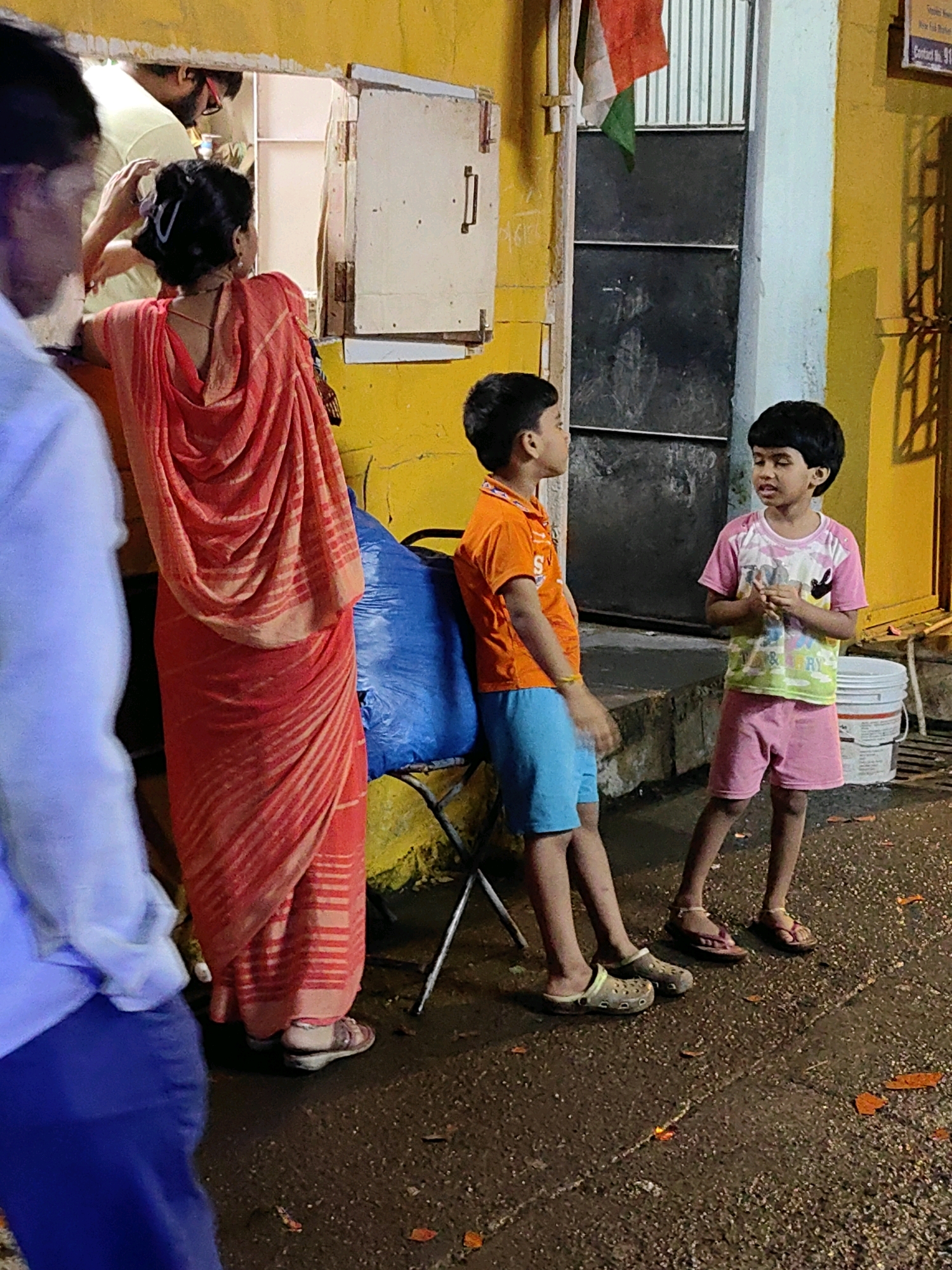 The image captures a street scene in Mumbai, India, likely at night or during overcast weather, given the dim lighting and wet ground. A woman in a vibrant orange sari with a striped pattern is the central figure in the mid-ground, interacting with a vendor through a window in a bright yellow building. Her sari is draped over her shoulders and torso, with the fabric pleated and flowing.
To the right of the woman, two young boys are standing on the wet pavement. The boy on the left wears an orange t-shirt and blue shorts, with a yellow bracelet on his left wrist. He is looking towards the boy on the right. The boy on the right wears a pink t-shirt with a colorful graphic and pink shorts. He is looking towards the left and appears to be speaking. Both boys are wearing casual footwear.
In the background, the yellow building has a dark metal door or shutter and a small Indian flag is visible, attached to a pole. The ground is a dark, textured surface, possibly asphalt or concrete, with scattered debris. A large blue bag is propped against the building near the woman. The overall impression is of an everyday scene in a bustling urban environment, with a focus on the human interactions taking place.