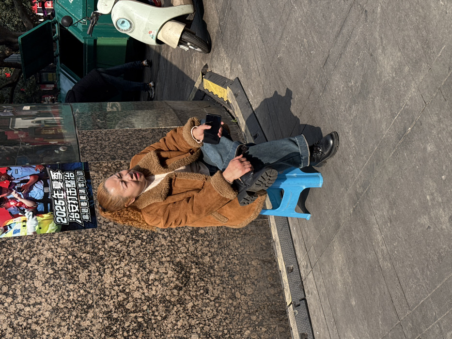 A person sits on a blue stool outdoors, wearing a brown shearling jacket and jeans. They hold a black smartphone in one hand and a black boot in the other. A poster with Chinese characters and images of police officers is to their left. A green dumpster and a white scooter are in the background. The scene is brightly lit, casting shadows on the paved surface.