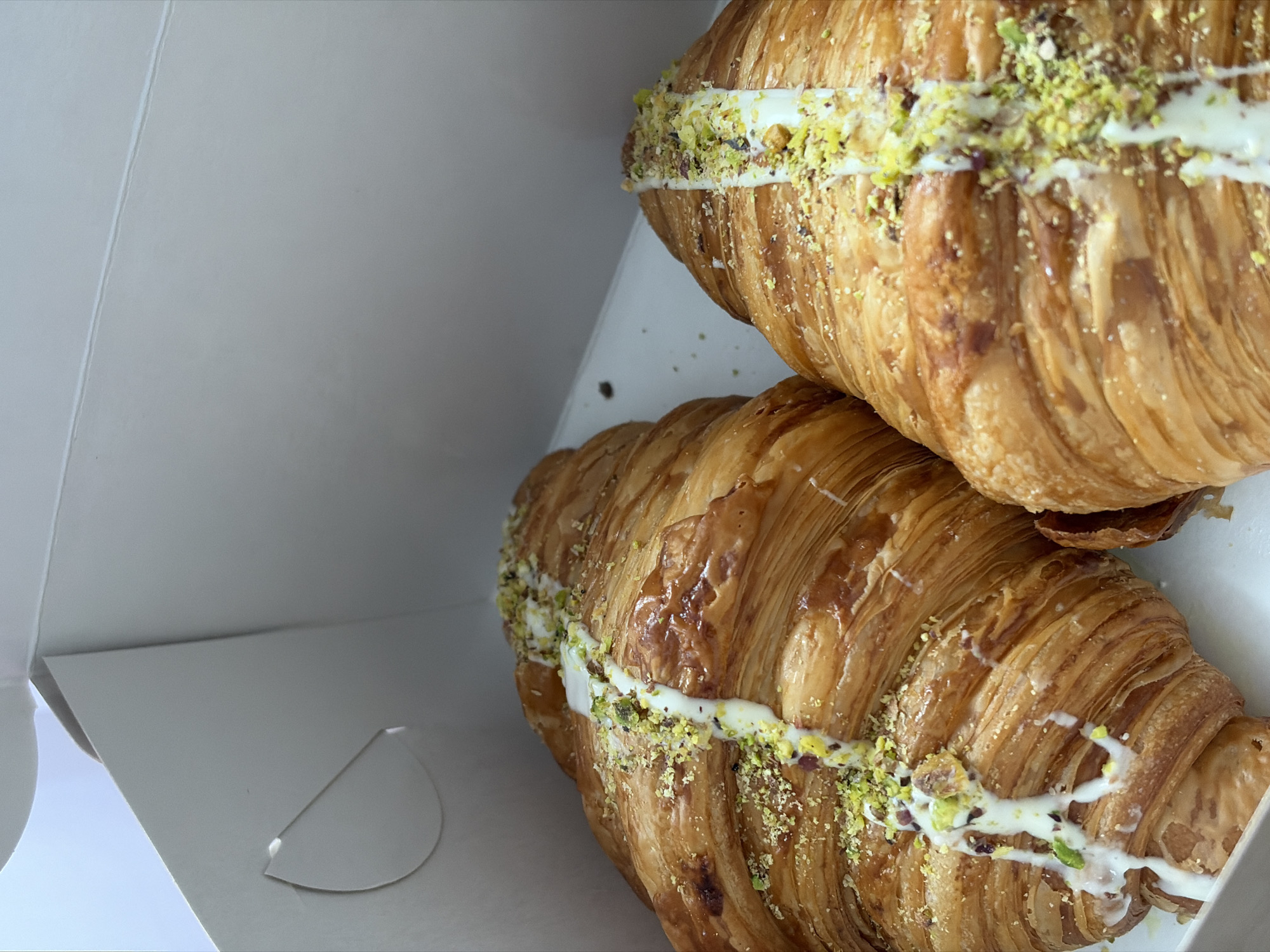 The image shows two filled croissants in a white cardboard box. The croissants are golden brown and glazed. They are filled with a white cream or frosting, and sprinkled with chopped pistachios. The box appears to be new and unused. The image does not contain information about a location in Votkinsk, Russia, or any people or interactions.