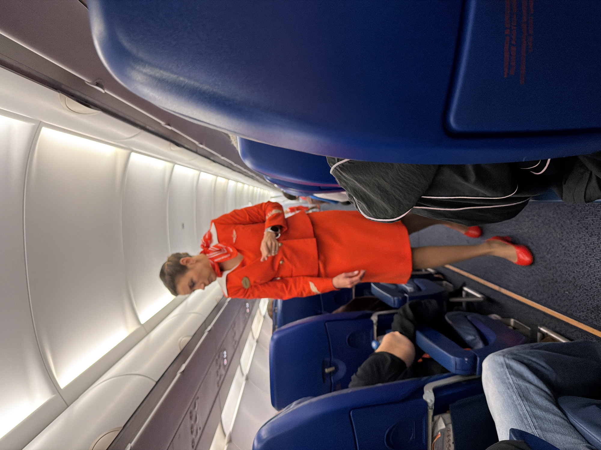 The image depicts the interior of a commercial airplane cabin, specifically the aisleway between rows of blue seats. A flight attendant, wearing an orange uniform including a skirt, jacket, scarf, and red heels, is the primary subject, standing in the aisle and looking downwards. The cabin environment is characterized by overhead luggage bins, lighting fixtures on the ceiling, and seatbacks of passengers. The aisle is carpeted in blue and marked with a yellow line. The scene appears to be in Saint Petersburg, Russia, given the context of the query, but this is not visually verifiable from the image itself.