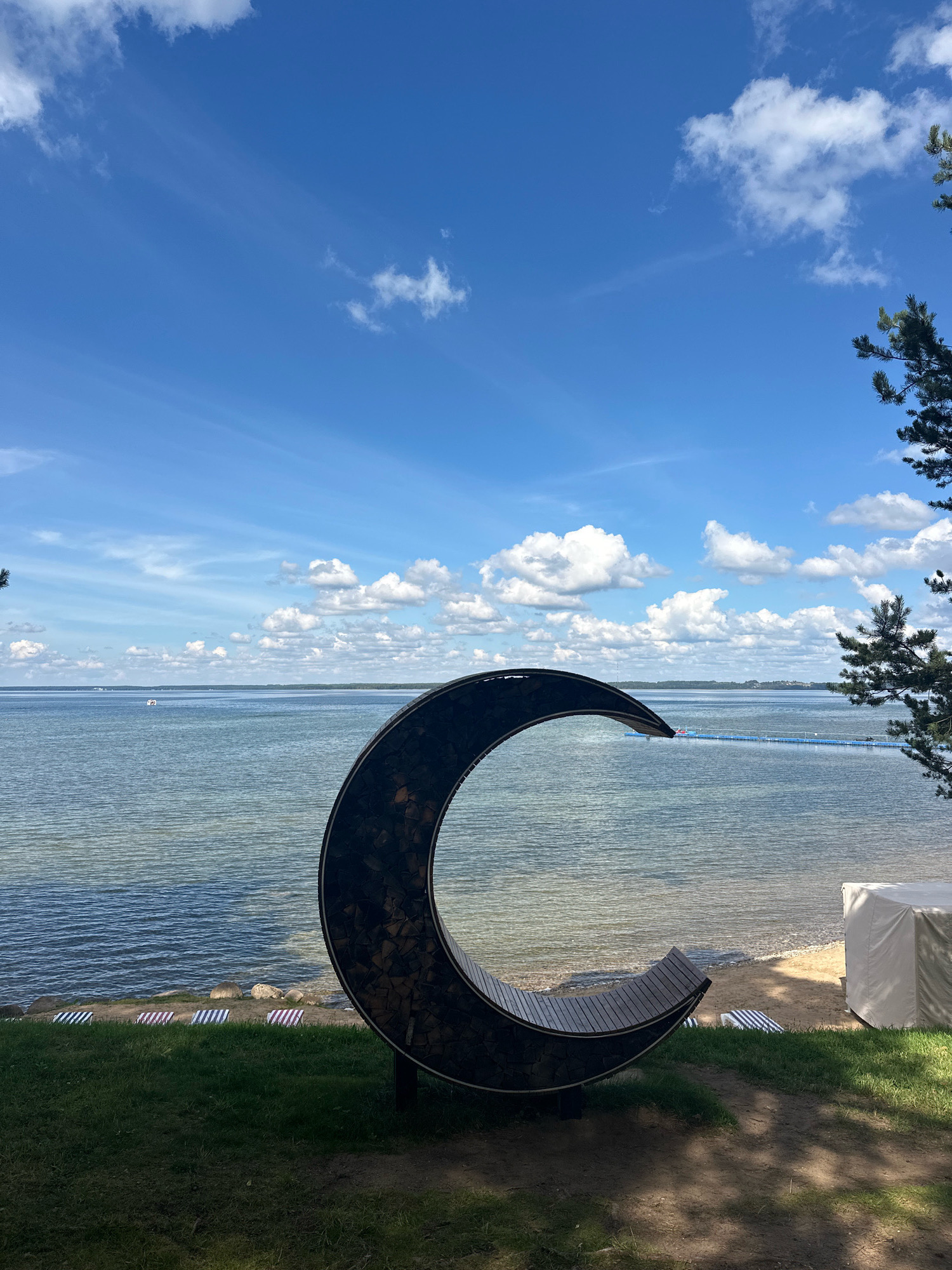 A crescent-shaped bench with a dark, textured outer shell and wooden slats for seating is positioned in the foreground on a grassy area overlooking a wide body of water. The water is calm, with a visible shoreline and a faint blue floating structure in the distance. The sky above is a vibrant blue, dotted with fluffy white clouds. To the right of the bench, a portion of a pine tree with green needles is visible, and further to the right, a white covered structure sits on the sandy beach. Several striped lounge chairs are placed on the beach near the water's edge. The overall scene suggests a serene outdoor setting, possibly a park or resort area.