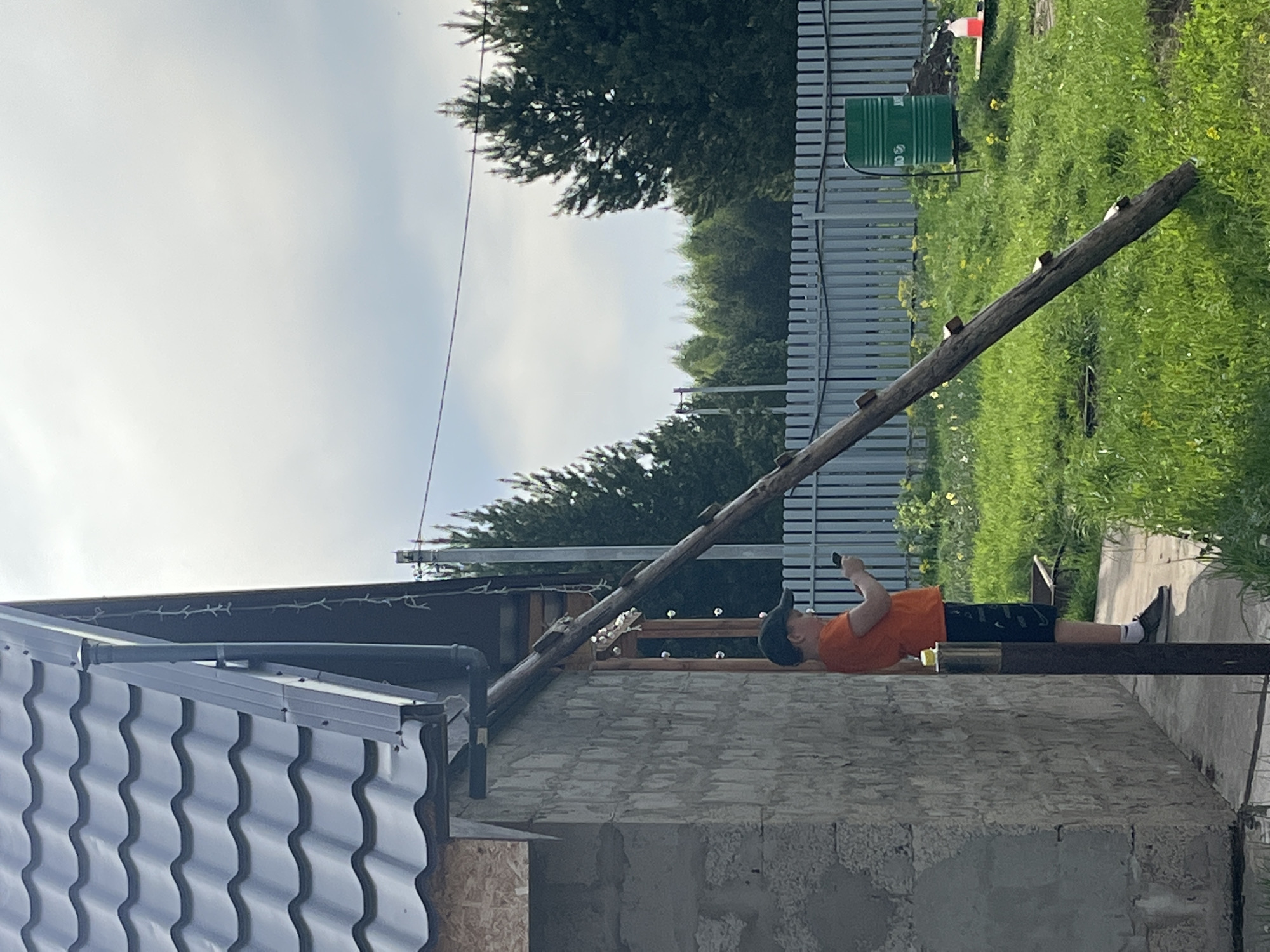 The photograph shows a scene in Гаревая, Russia. A young person wearing a black baseball cap, orange t-shirt, and black shorts is lying on a horizontal, roughly hewn wooden beam. The beam extends from the top of a partially constructed building with a corrugated metal roof. A wooden ladder is leaning against the building, leading up to the beam. The person is holding a mobile device, presumably a smartphone, in their hand. Behind the building and the beam is a gray fence, a green barrel or container, and a grassy area with trees in the background. The sky is overcast.