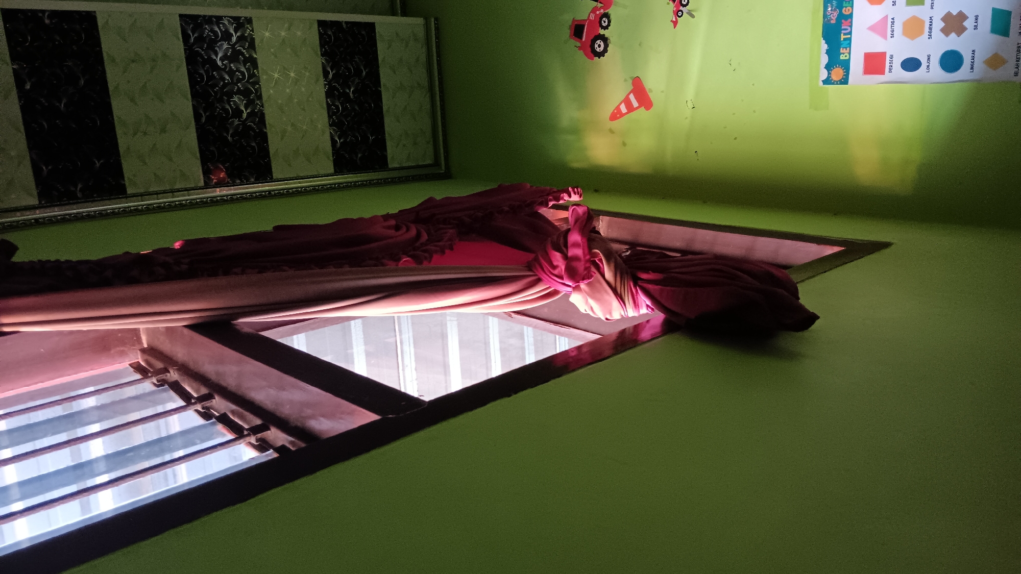 The image depicts an interior scene with a focus on a window and its surroundings. The window is partially obscured by a draped, pinkish-red curtain that is tied back with a knot. The window frame appears to be dark brown, and horizontal blinds are visible through the glass. The walls of the room are painted a bright green. The ceiling has a decorative pattern of black and green stripes. On the green wall, there are several small, colorful objects, including a red toy truck, a red traffic cone, and a poster displaying various geometric shapes with their names in Indonesian. The overall impression is that of a room, possibly a child's room, in Indonesia, given the text on the poster. There are no people visible in the image.