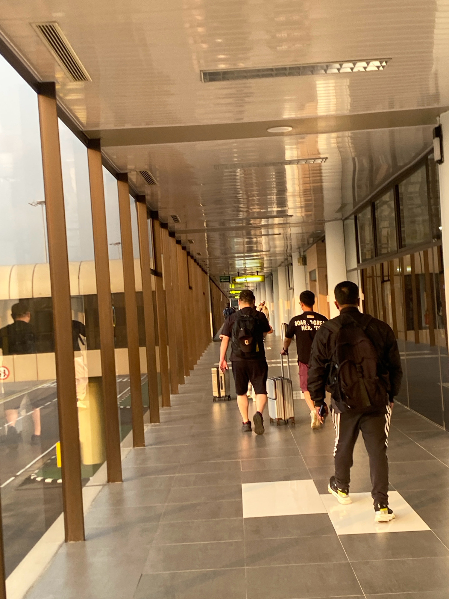 The image captures a view from an interior walkway at Hang Nadim International Airport (BTH) in Batam, Indonesia. The walkway features a modern, clean design with a tiled floor and a reflective ceiling with linear lighting and air vents. A row of vertical, light brown wooden beams lines the left side, separating the walkway from a large glass window offering a glimpse of the outside, which appears to be an airport tarmac. Through the glass, a segment of a white airport structure or vehicle is visible, along with some ground markings that suggest a runway or boarding area.
Several individuals are walking along the walkway, all facing away from the camera and towards the end of the corridor. The most prominent subjects are three men in the foreground and mid-ground. The man closest to the camera is wearing a black hooded jacket over a t-shirt, black pants with white stripes, and white and yellow athletic shoes. He is carrying a black backpack. To his left, a man is wearing a black t-shirt with white text that reads "SOAR GREATER HEIGHTS" and maroon shorts. He is pulling a silver rolling suitcase. Further ahead and to the left, another man is wearing a black t-shirt and black shorts, also with a black backpack, and he is walking alongside another rolling suitcase. There are other individuals further down the walkway, less distinct due to distance and focus. The lighting suggests it might be late afternoon or early evening, with warm, diffused light entering from the left. The overall atmosphere is one of transit and movement within an airport environment.