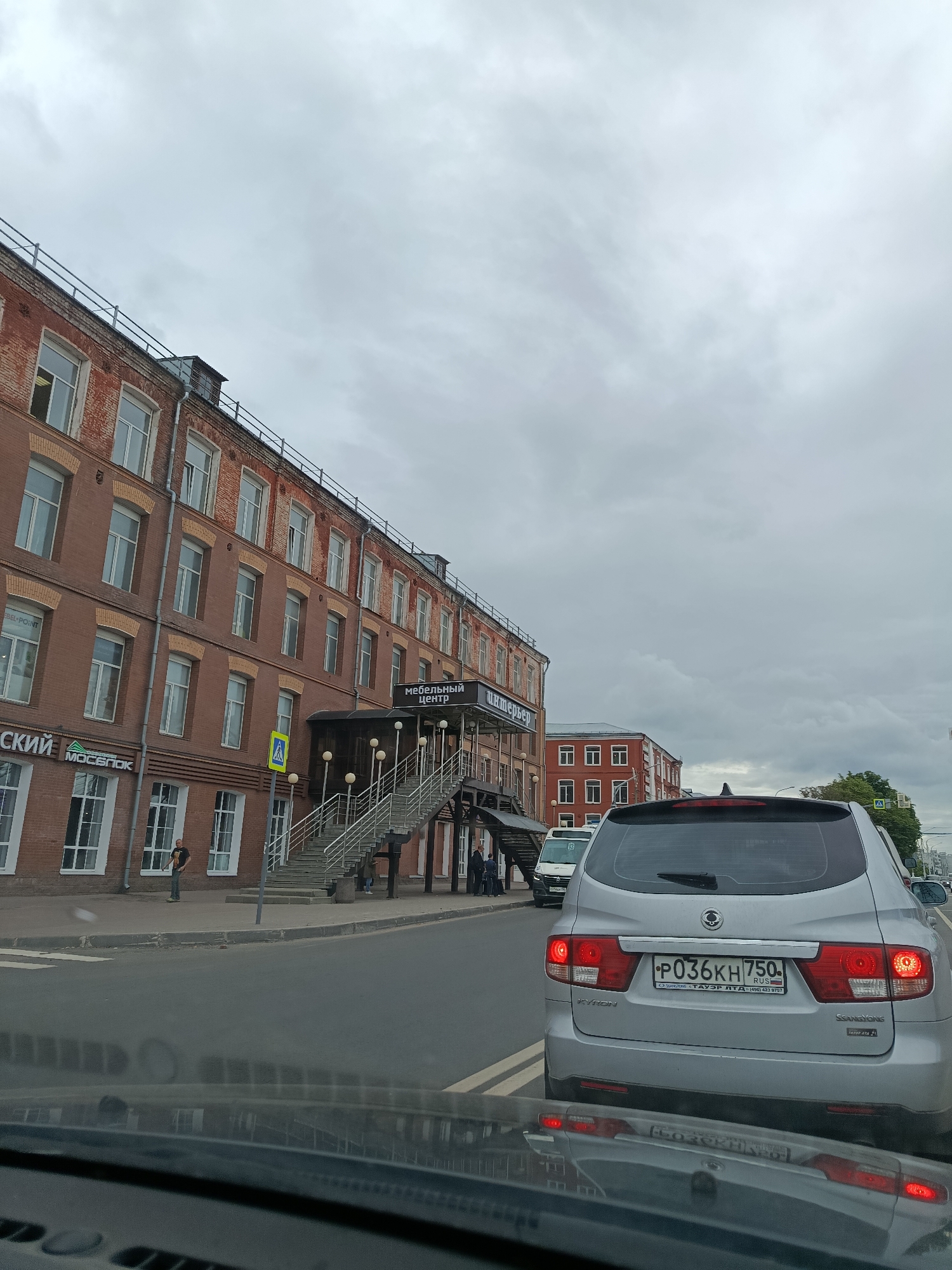This media file captures a street scene in Orekhovo-Zuyevo, Russia, on an overcast day. The primary subject is a large, multi-story brick building on the left, appearing to be a commercial or industrial establishment. The building features numerous windows and an external staircase leading to an entrance marked with Cyrillic text that translates to "Furniture Center." Below this entrance, a sign reads "BEL-POINT" and another sign displays "MOSBLOK" in Cyrillic. A yellow pedestrian crossing sign is visible on the sidewalk.
In the foreground, the rear of a silver SsangYong Kyron SUV is prominently displayed, its brake lights illuminated. The license plate is visible, indicating it's a Russian vehicle with the registration P036KH 750. The car is stopped on a road, suggesting traffic or a pause in movement. A white van with the number "13" on its side is visible ahead of the SUV.
Several individuals are present in the scene: one person stands on the sidewalk near the brick building, and a few others are seen on the street near the entrance of the furniture center. The sky is heavily clouded, indicating a gloomy or possibly rainy weather condition, and the time of day appears to be daytime. The overall atmosphere is that of a typical urban street environment.
