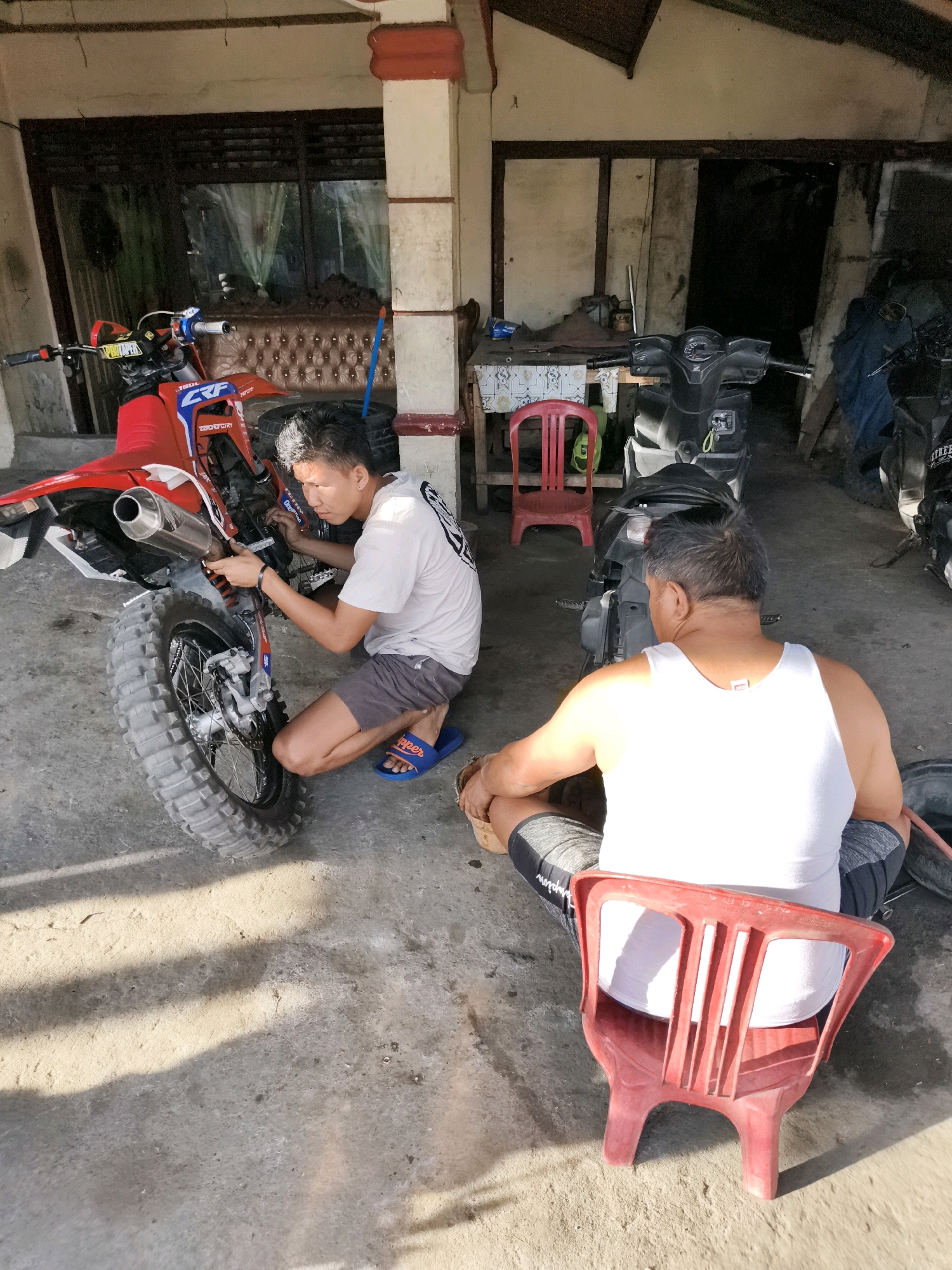 Two men are working on a red dirt bike in a covered outdoor area, possibly a garage or workshop, in Tombatu, Indonesia. The man on the left, wearing a white t-shirt and grey shorts, is kneeling and appears to be adjusting or repairing the rear wheel of the dirt bike. He has blue flip-flops on his feet. The man on the right, wearing a white tank top and grey shorts with black stripes, is sitting on a red plastic chair and facing away from the camera. He is looking towards the dirt bike. The dirt bike is red with white accents and has a large knobby tire. There are other motorcycles and what appears to be a table with items on it in the background. The ground is concrete and there are shadows cast from sunlight. The environment suggests a typical setting for vehicle maintenance or repair in a warm climate.