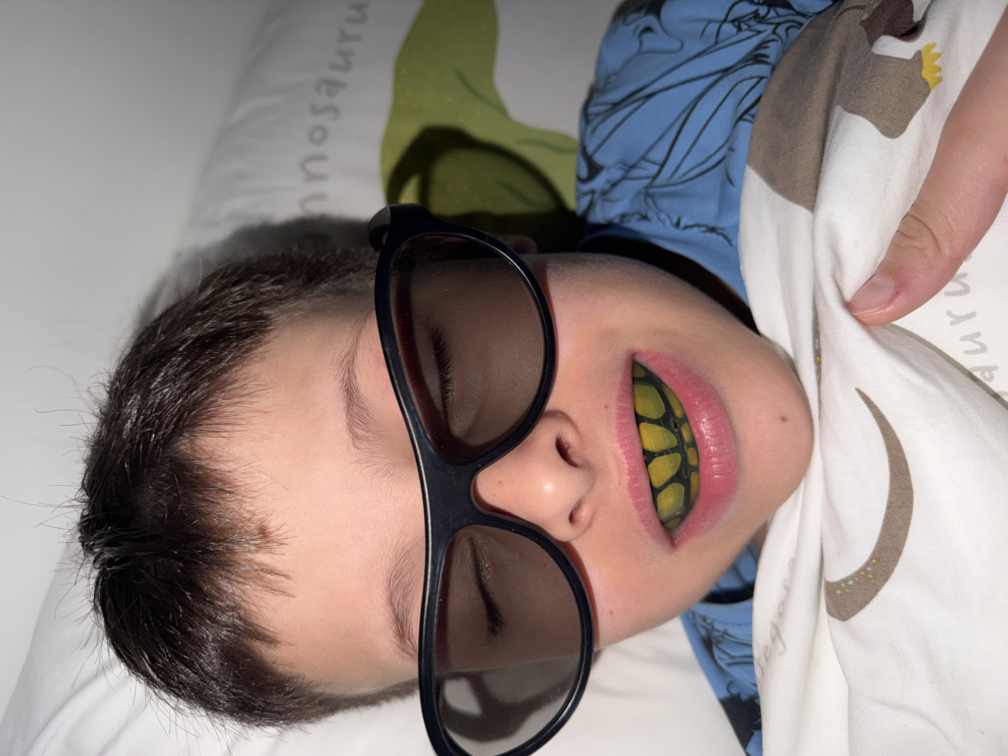 A young boy lies on a white pillow with a dinosaur print. He is wearing 3D glasses and a yellow and black mouthguard. He is also wearing a blue and black shirt. His hand is holding the white dinosaur-printed pillow. The location is unknown, and there is no evidence to suggest the image was taken in Votkinsk, Russia.