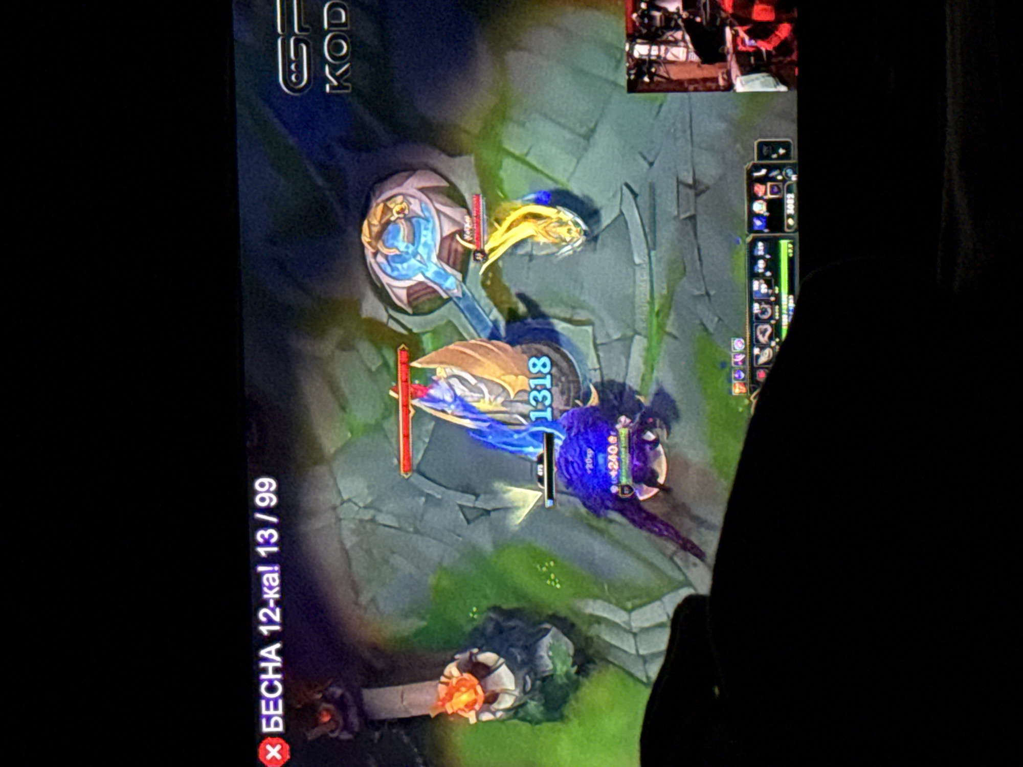The image shows a video game being played on a screen. The game appears to be a multiplayer online battle arena (MOBA) game, possibly League of Legends. Characters are engaging in combat, as evidenced by the health bars and damage numbers. In the top right corner, there is a smaller screen possibly showcasing the user or other content. A blurred human figure is partially visible on the right side of the image. Text on the screen indicates game-related information, such as "БЕСНА 12-ка! 13/99", damage numbers, and other in-game stats.