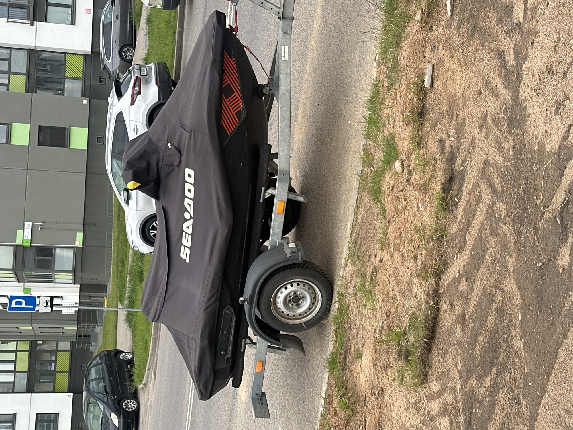 A black Sea-Doo personal watercraft on a trailer is parked on the side of a road in Бараўлянскі сельскі Савет, Belarus. The watercraft is covered with a gray cover displaying the Sea-Doo logo in white. The trailer has a single wheel and gray frame. The road is asphalt with a dirt and grass shoulder. Cars are parked along the road near an apartment building with green and gray panels. A blue parking sign is visible next to the road.