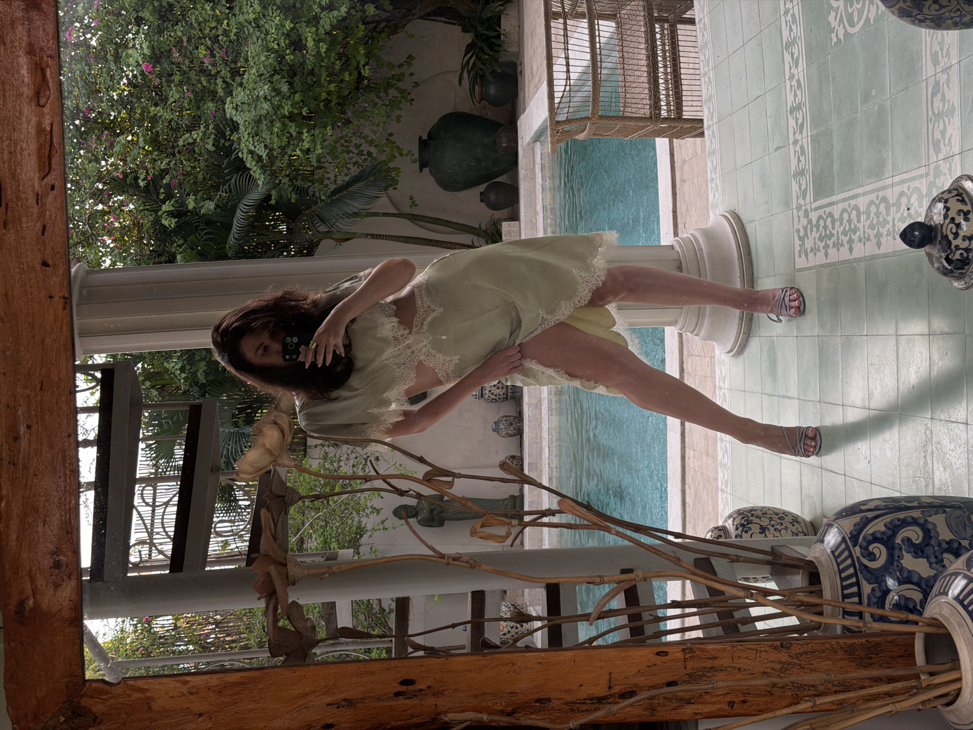 A woman stands in front of a mirror in what appears to be an outdoor courtyard in North Kuta, Indonesia. The woman is wearing a light green slip dress with lace detailing and is holding a smartphone to take a mirror selfie. She is wearing strappy heeled sandals. Behind her, visible in the mirror's reflection, is a swimming pool with clear blue water, surrounded by lush greenery and potted plants. A spiral staircase and a statue are also visible in the reflection, suggesting an architectural feature of the property. The floor is tiled in a light green and white pattern. A wicker chair is positioned near the pool. Decorative vases are placed around the courtyard. The overall impression is one of a tropical, luxurious setting.